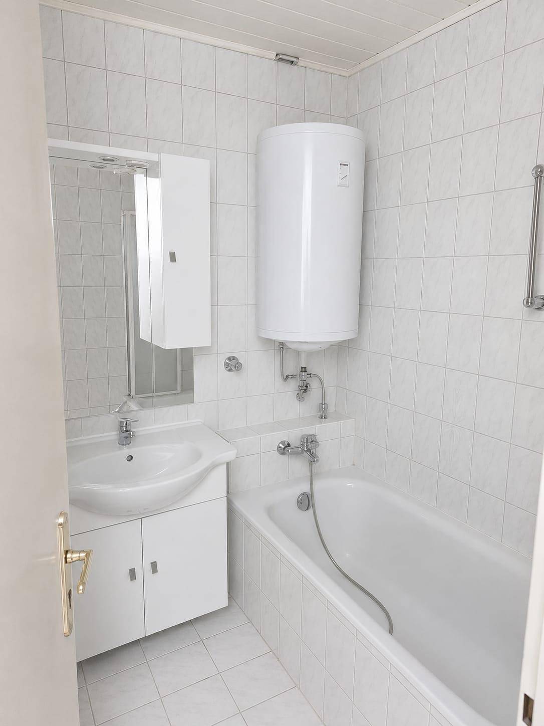 Helles Badezimmer mit Badewanne, Waschbecken und weiß gefliesten Wänden, ausgestattet mit einem Boiler.
