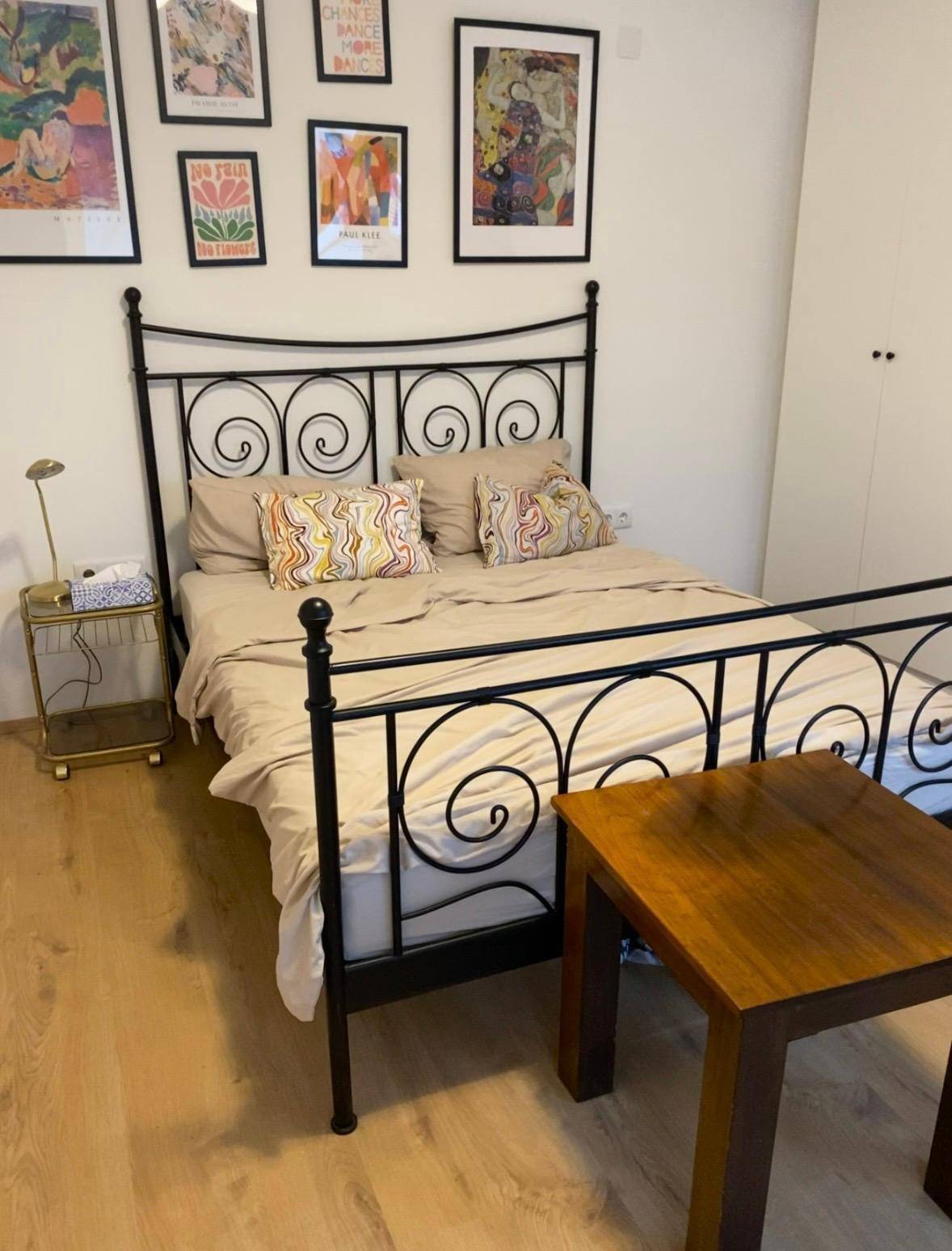 Schlafzimmer mit schmiedeeisernem Bettgestell, Holzboden und farbenfrohen Kunstwerken an der Wand.