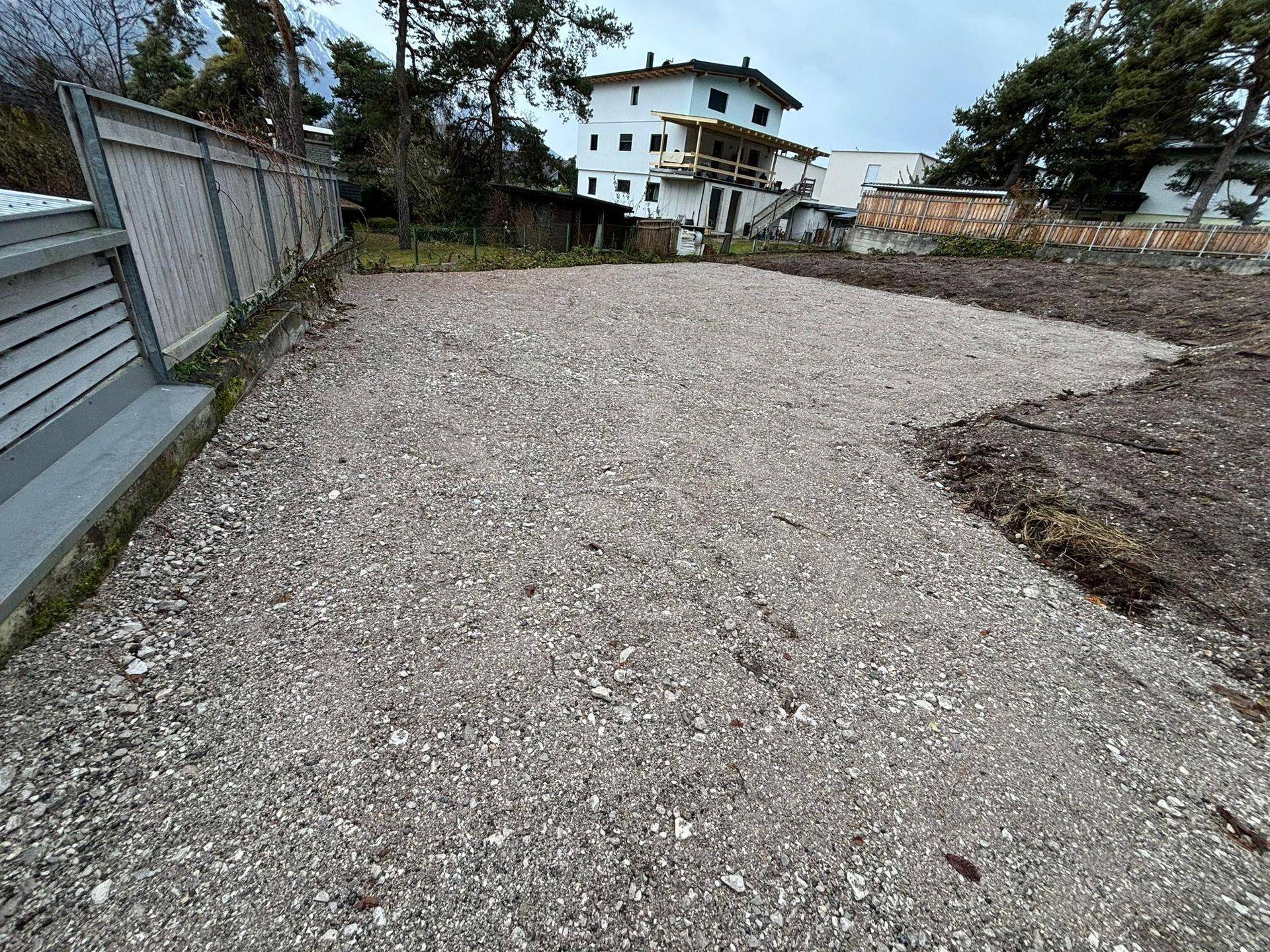 Weitläufige Schotterfläche neben einem Zaun, umgeben von Bäumen und einem Gebäude im Hintergrund.