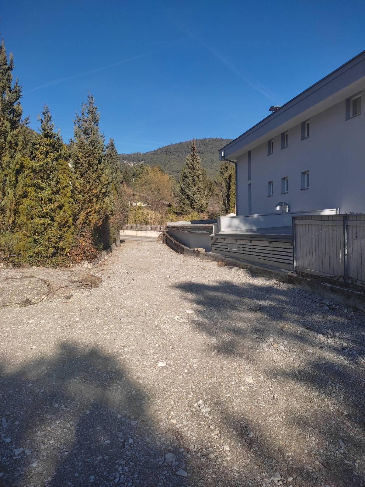 Sonnenbeschienener Schotterweg führt zu einem modernen Gebäude mit Bergen im Hintergrund unter blauem Himmel.