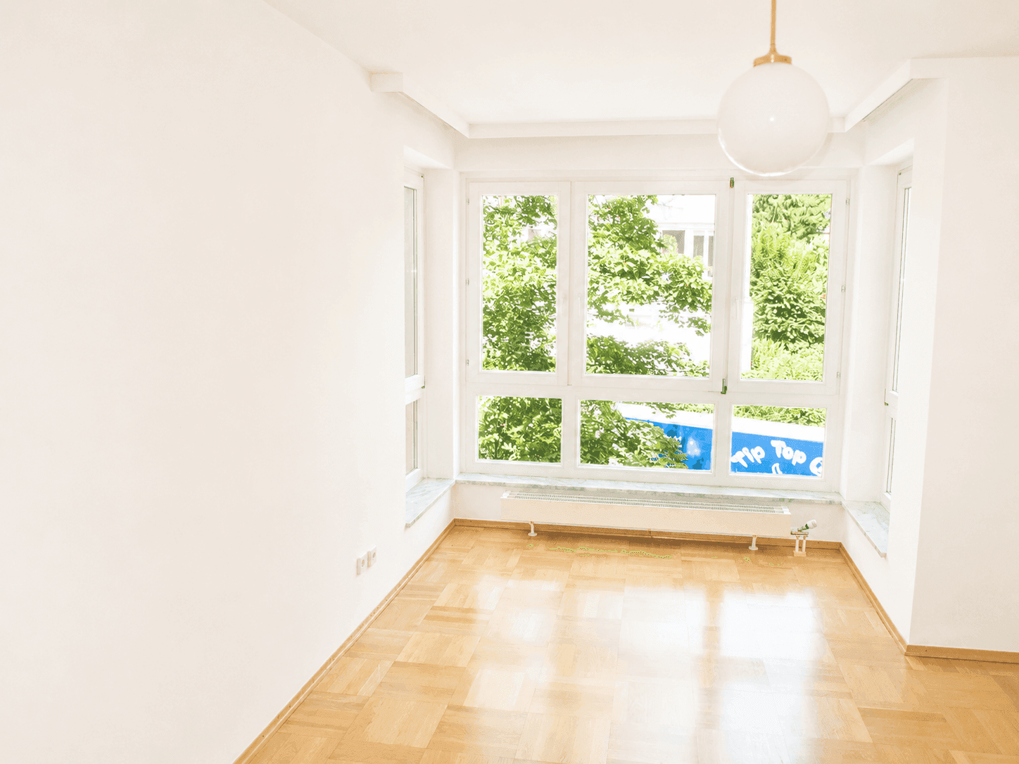 Heller Raum mit großen Fenstern, Parkettboden und Blick auf die umliegende grüne Umgebung.