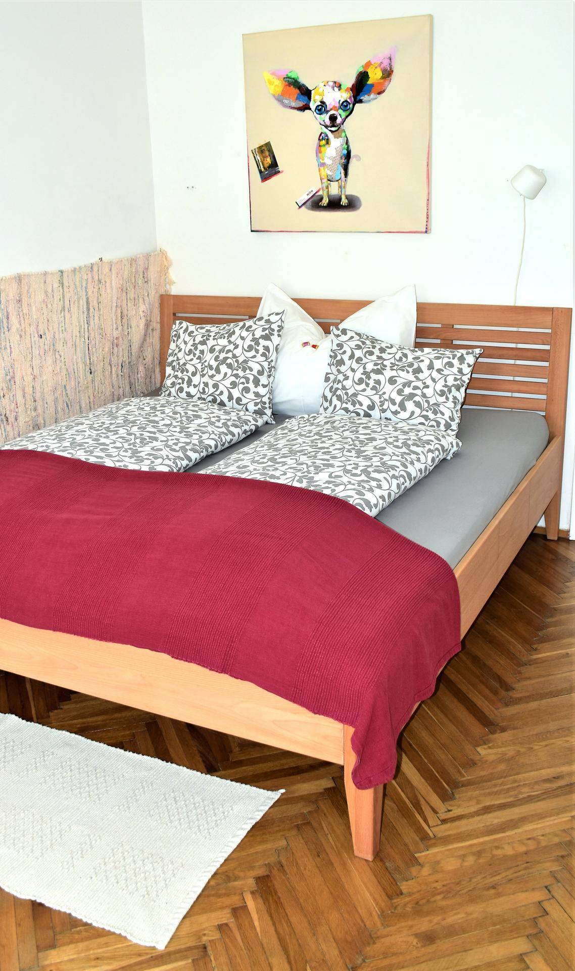 Schlafzimmer mit Holzbett, roter Decke und klassischem Fischgrätparkett, dekoriert mit einem farbenfrohen Bild.