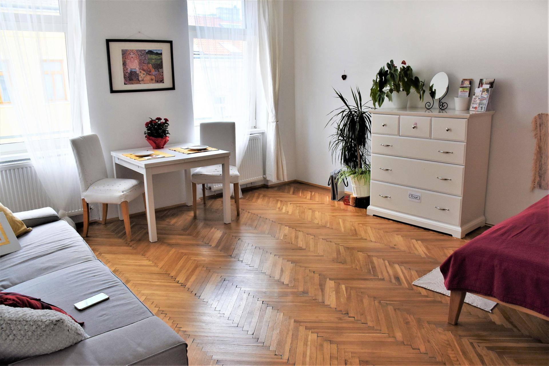 Heller offener Wohnbereich mit Sofa, Esstisch, Kommode und Blick auf das Schlafzimmer, mit Fischgrätparkett.