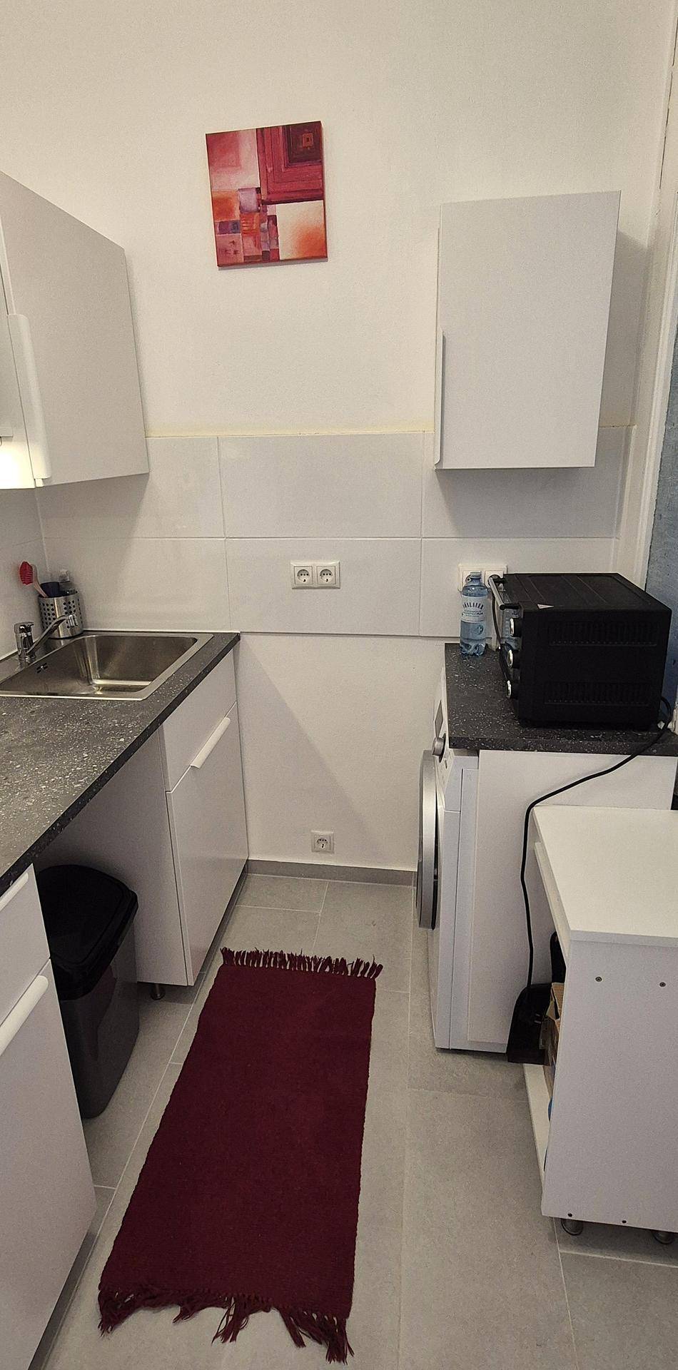 Funktionale Küchenzeile mit Waschmaschine, weißen Schränken und dunkler Arbeitsplatte, auf Fliesenboden.