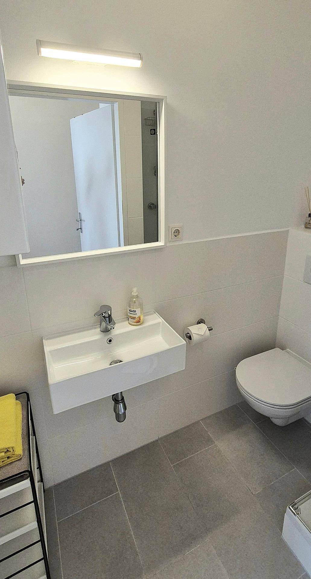 Modernes Badezimmer mit wandhängendem WC, Einzelwaschbecken und großem Spiegel, hell gefliest.