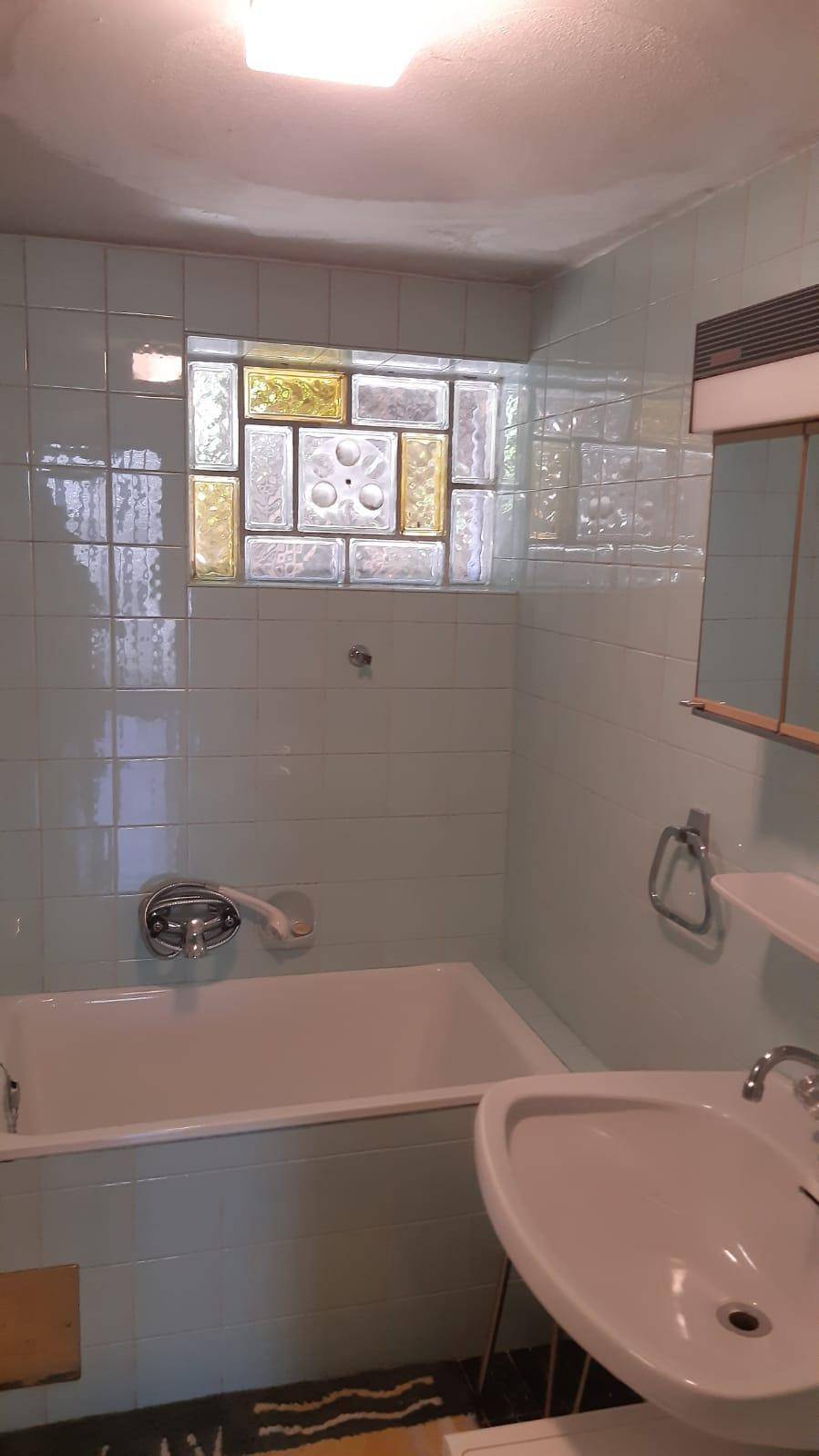 Badezimmer mit Badewanne, Waschbecken und einem Fenster aus Glasbausteinen, hell gefliest.