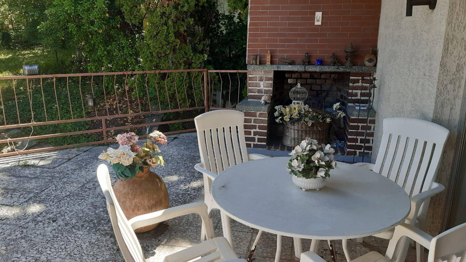 Gemütliche Terrasse mit Außenkamin und einfachen Gartenmöbeln, umgeben von viel Grün.