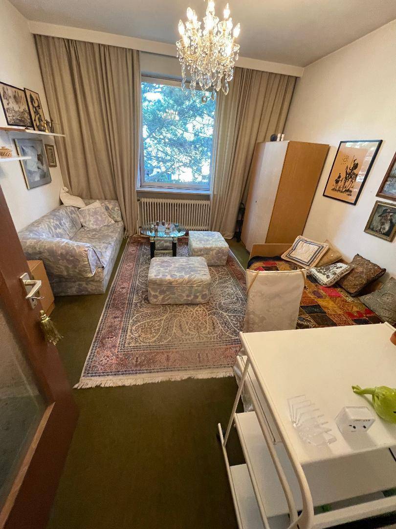 Helles Wohnzimmer mit Kronleuchter, Teppichboden und einem Fenster mit Blick ins Grüne.