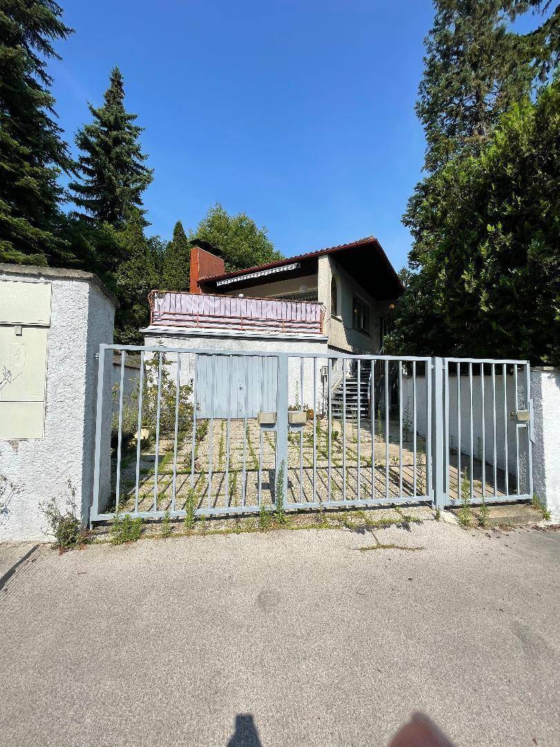 Eingangsbereich des Hauses mit Metalltor, gepflasterter Auffahrt und umgebenden Bäumen.