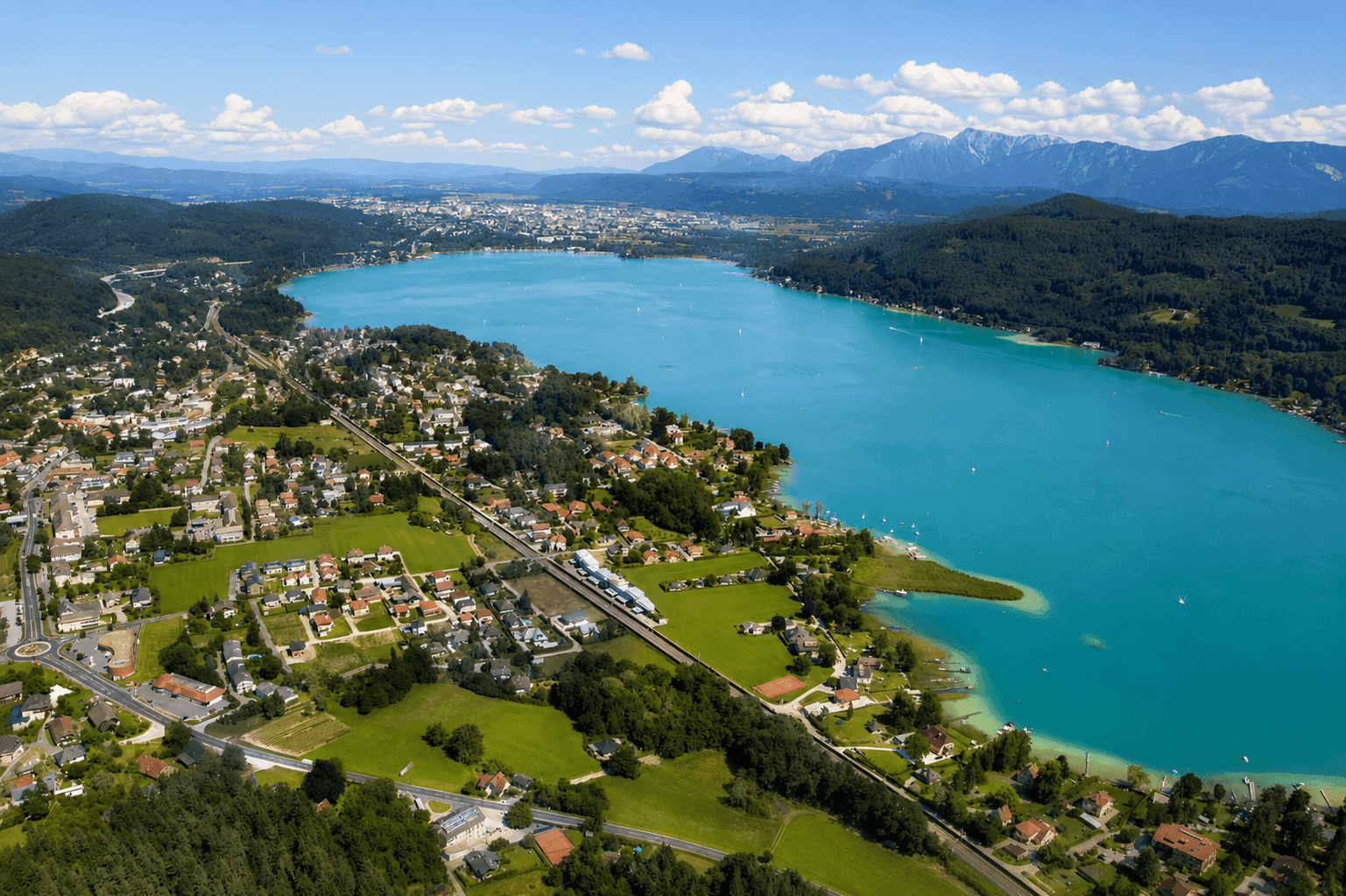 Luftaufnahme des türkisblauen Ossiacher Sees mit umliegenden Ortschaften und Bergen im Hintergrund.