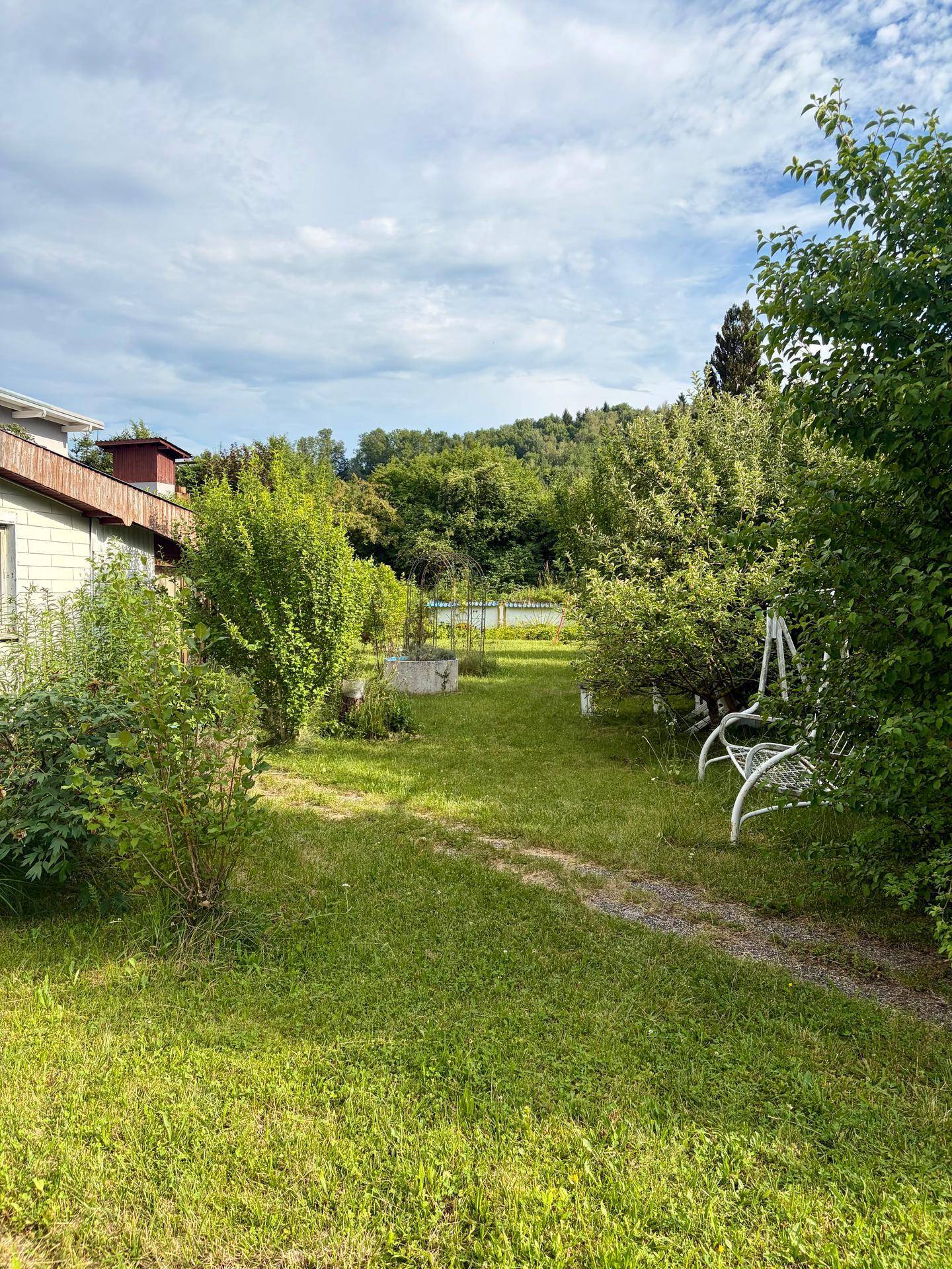 Weitläufiger grüner Garten mit einer Schaukelbank und üppiger Vegetation, umgeben von Bäumen und Hügeln.