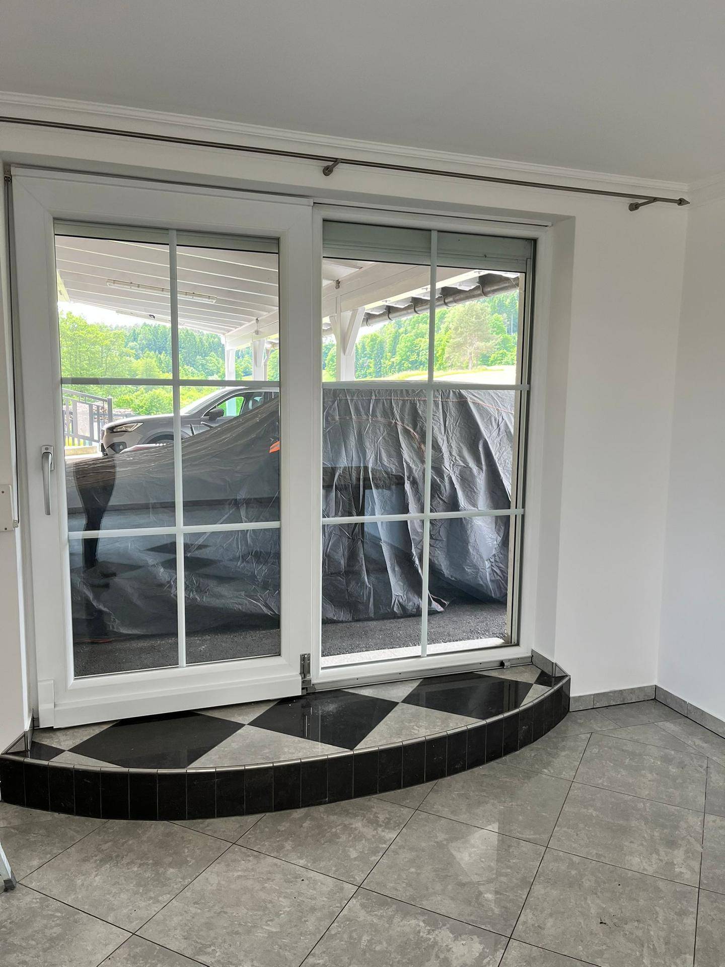 Große Fenstertür mit Blick auf einen überdachten Parkplatz und grüne Umgebung.