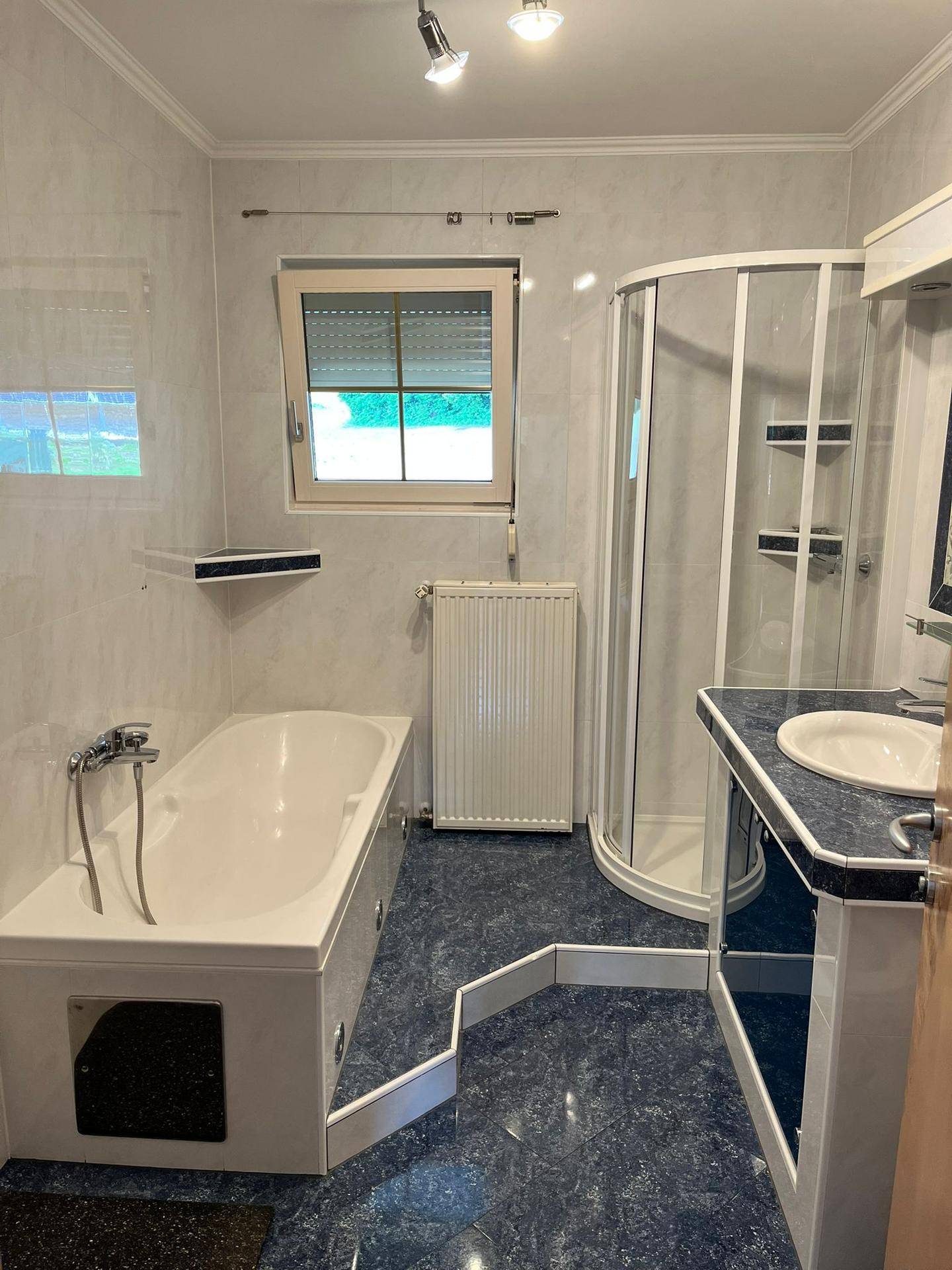 Badezimmer mit Badewanne, Duschkabine, Fenster und blauen Akzenten auf dem Boden und den Möbeln.