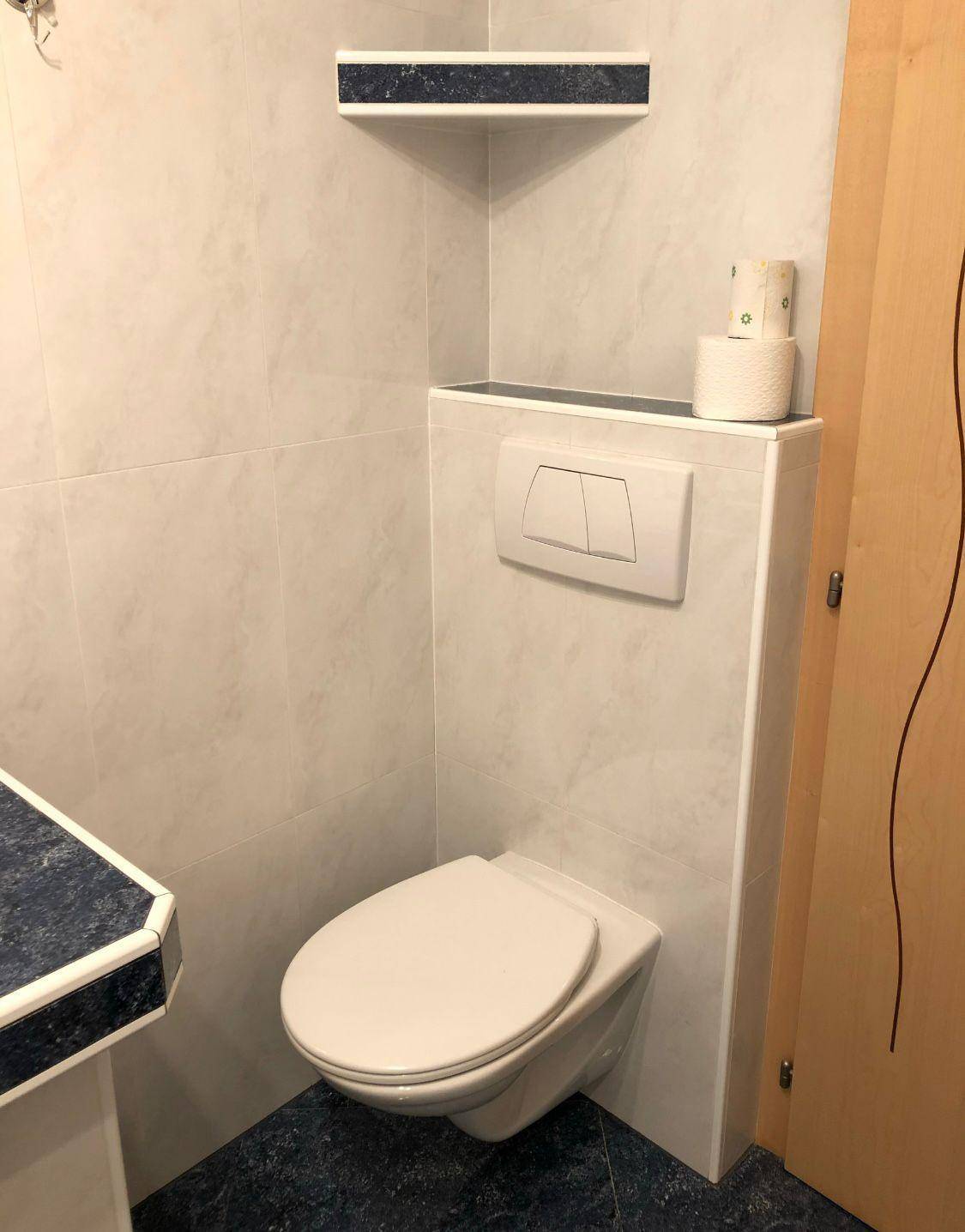Wandhängende Toilette mit Spülkasten und blauen Akzenten in einem gefliesten Badezimmer.