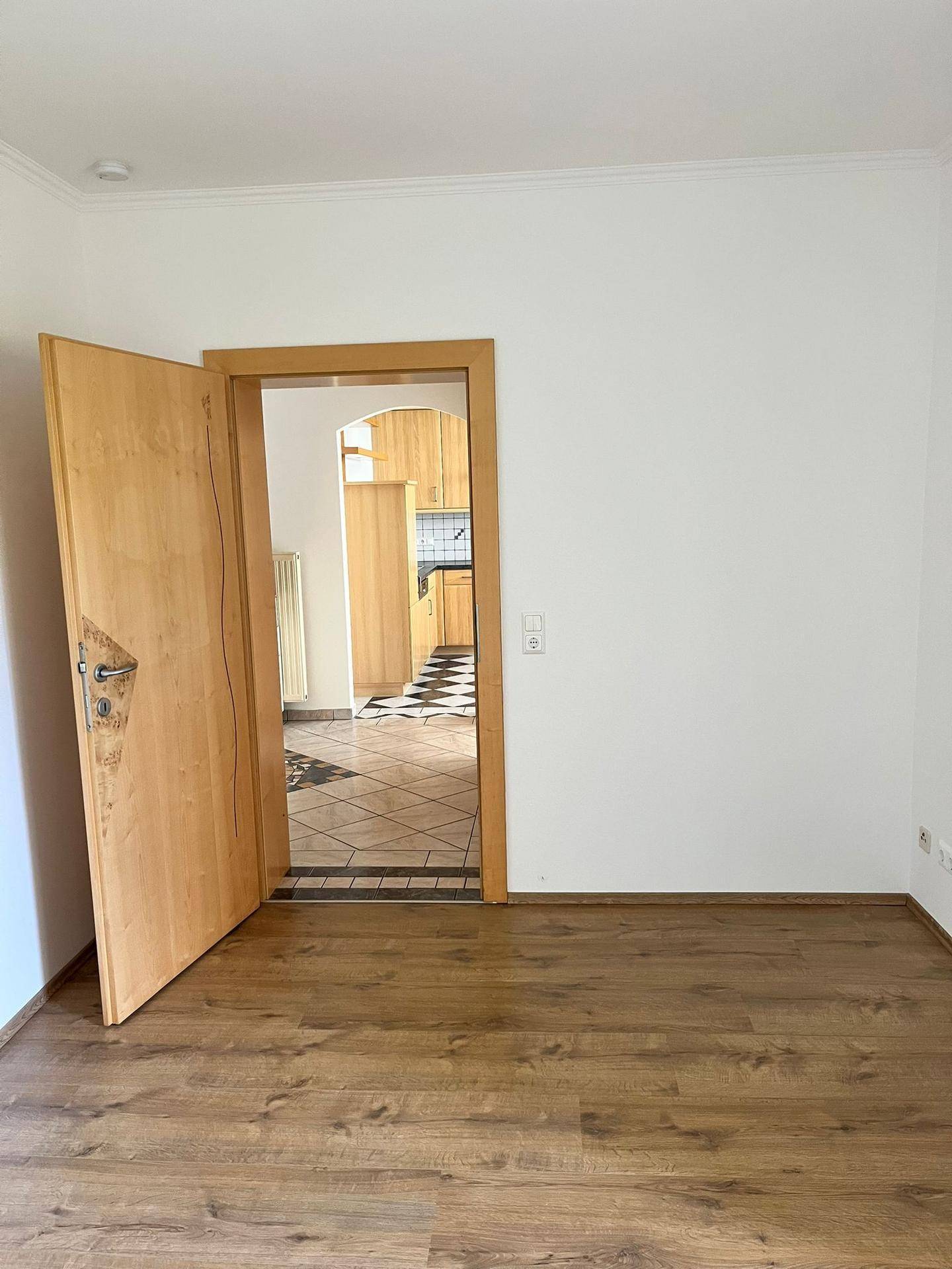 Heller Raum mit Holzboden und Blick durch eine offene Tür in die angrenzende Küche.