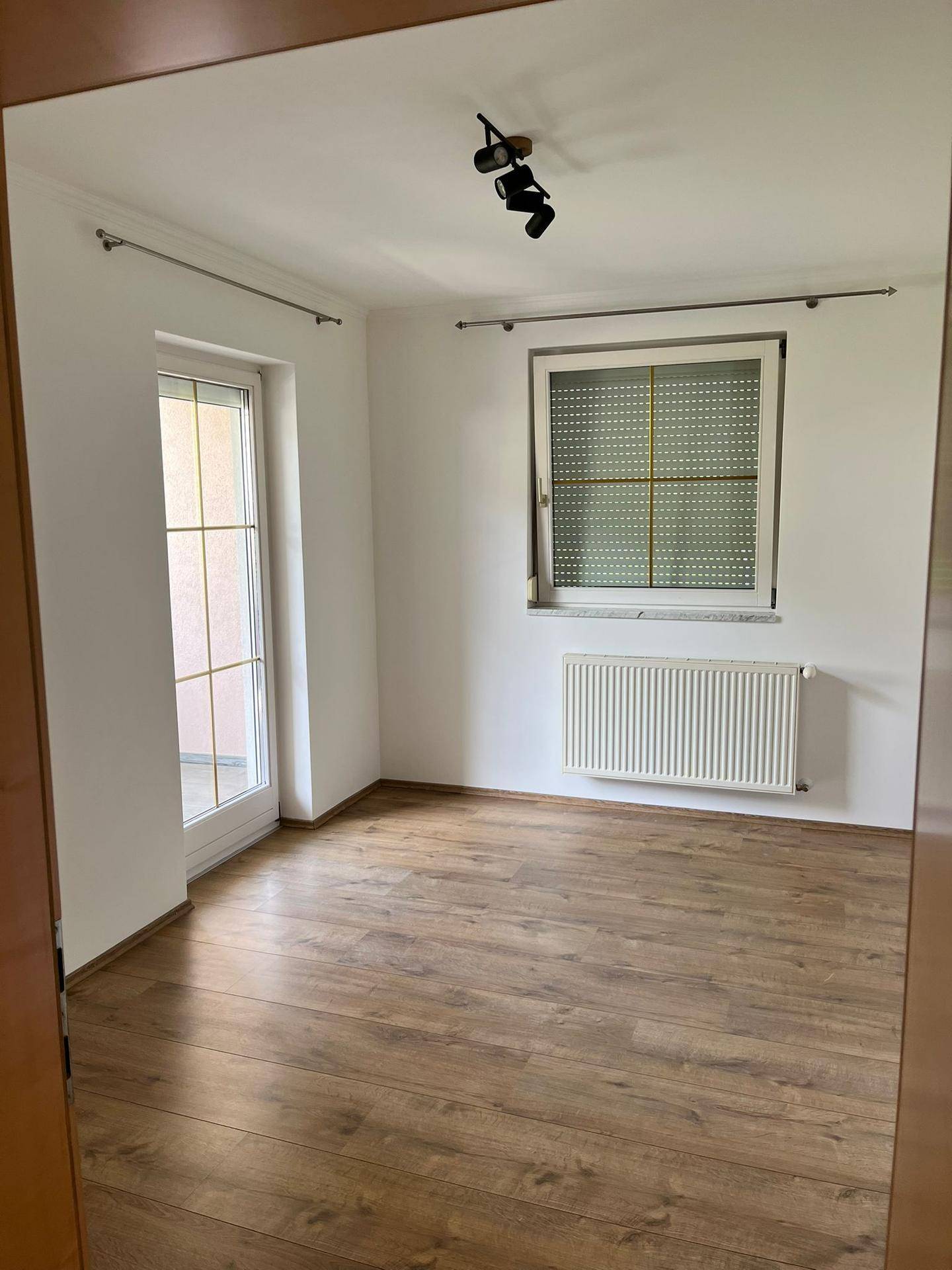 Heller Raum mit Laminatboden, Fenster und Zugang zu einem französischen Balkon.