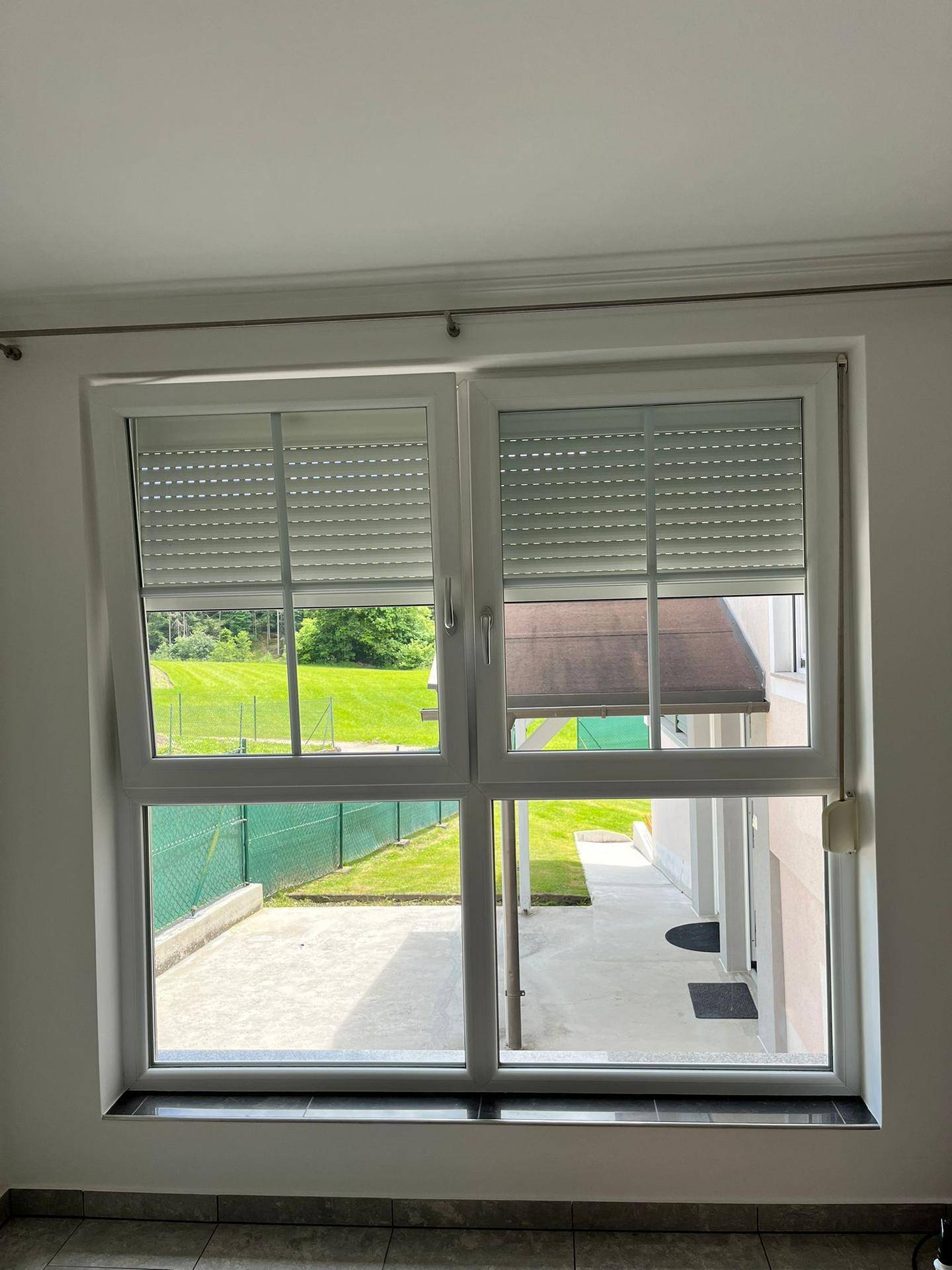 Großes Fenster mit Rollläden bietet einen weiten Blick auf grüne Wiesen und den Gartenbereich.