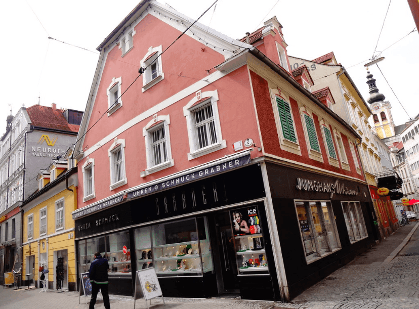 Traditionelles Gebäude mit rosa Fassade, grünen Fensterläden und Geschäften im Erdgeschoss.