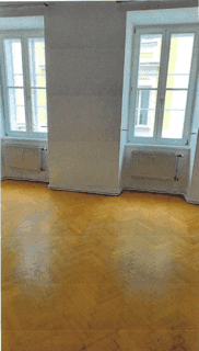 Heller Raum mit Holzboden und zwei hohen Fenstern, die viel Tageslicht hereinlassen.