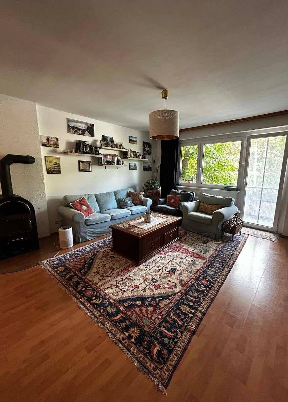 Helles Wohnzimmer mit Kaminofen, Sofa und Zugang zum Balkon, mit Holzboden und Teppich.