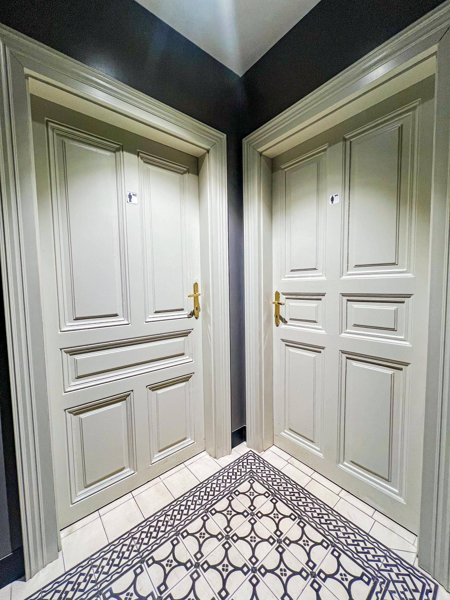 Flur mit zwei eleganten Türen, die zu den Toiletten führen, und einem gemusterten Fliesenboden.