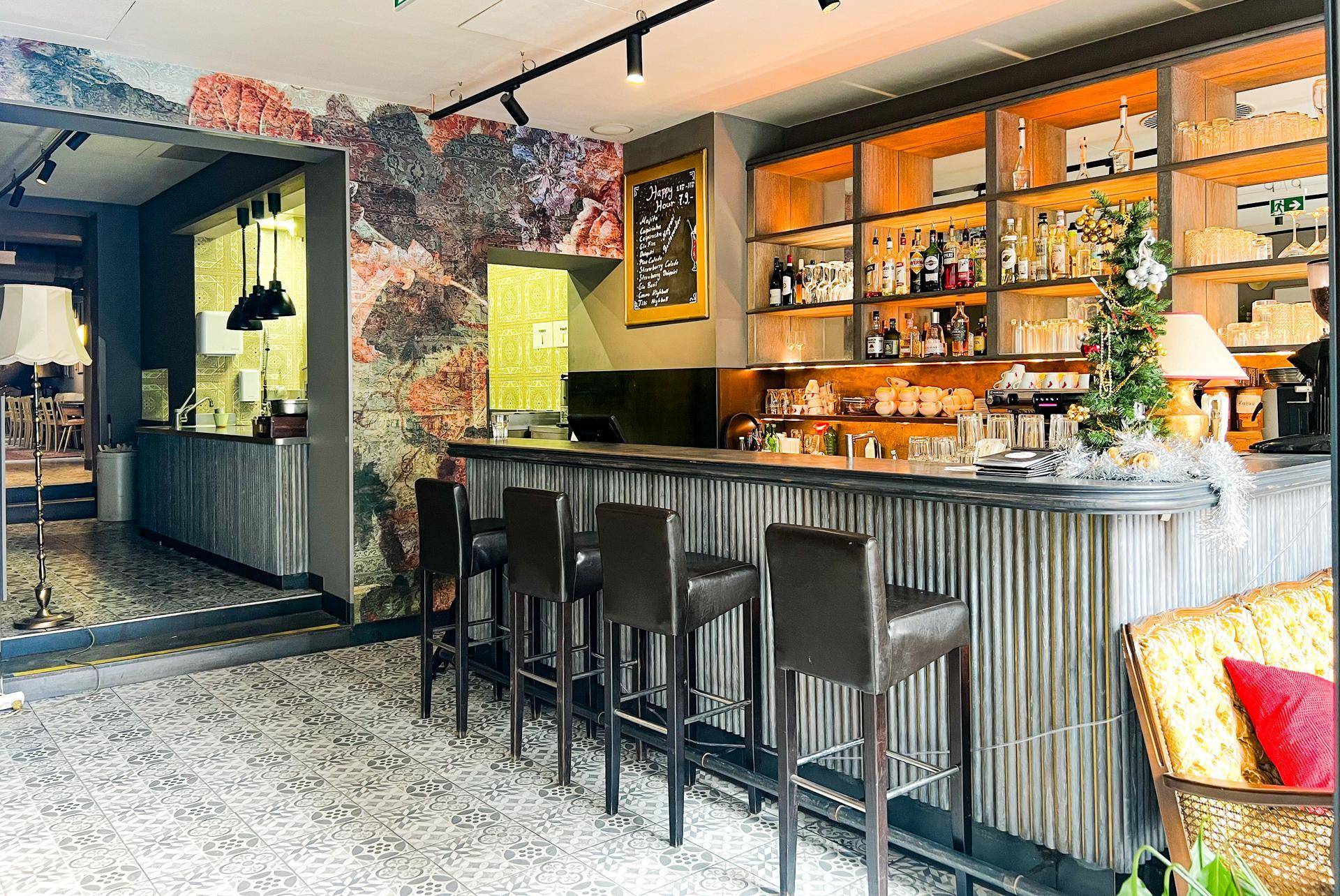 Stilvolle Bar mit dunklen Hockern, Holzregalen und einer dekorativen Wandtapete im Hintergrund.