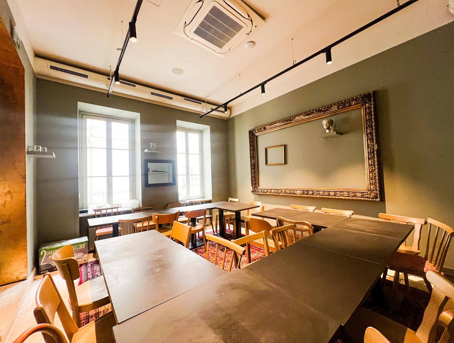 Heller Gastraum mit mehreren Tischen und Stühlen, großen Fenstern und einer schlichten Wandgestaltung.