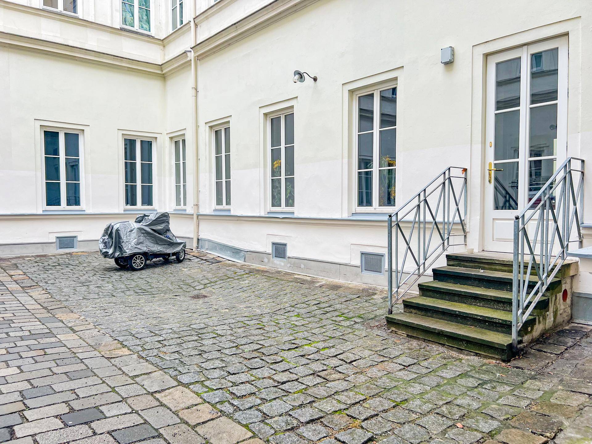 Innenhof mit Kopfsteinpflaster, weißen Fassaden, großen Fenstern und einer Treppe zum Eingang.