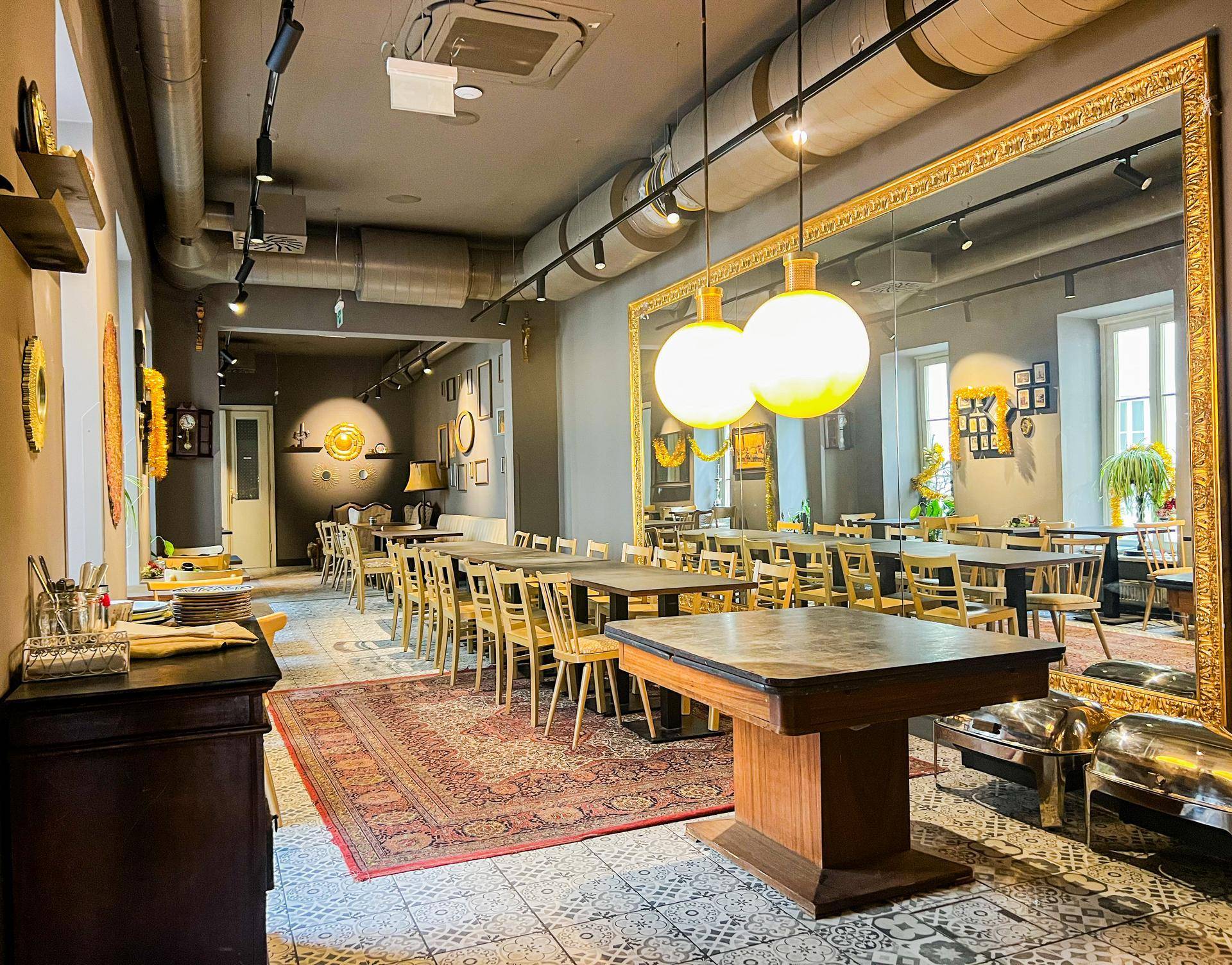 Großer Gastraum mit vielen Tischen, Stühlen, einem Teppich und einem imposanten Wandspiegel.