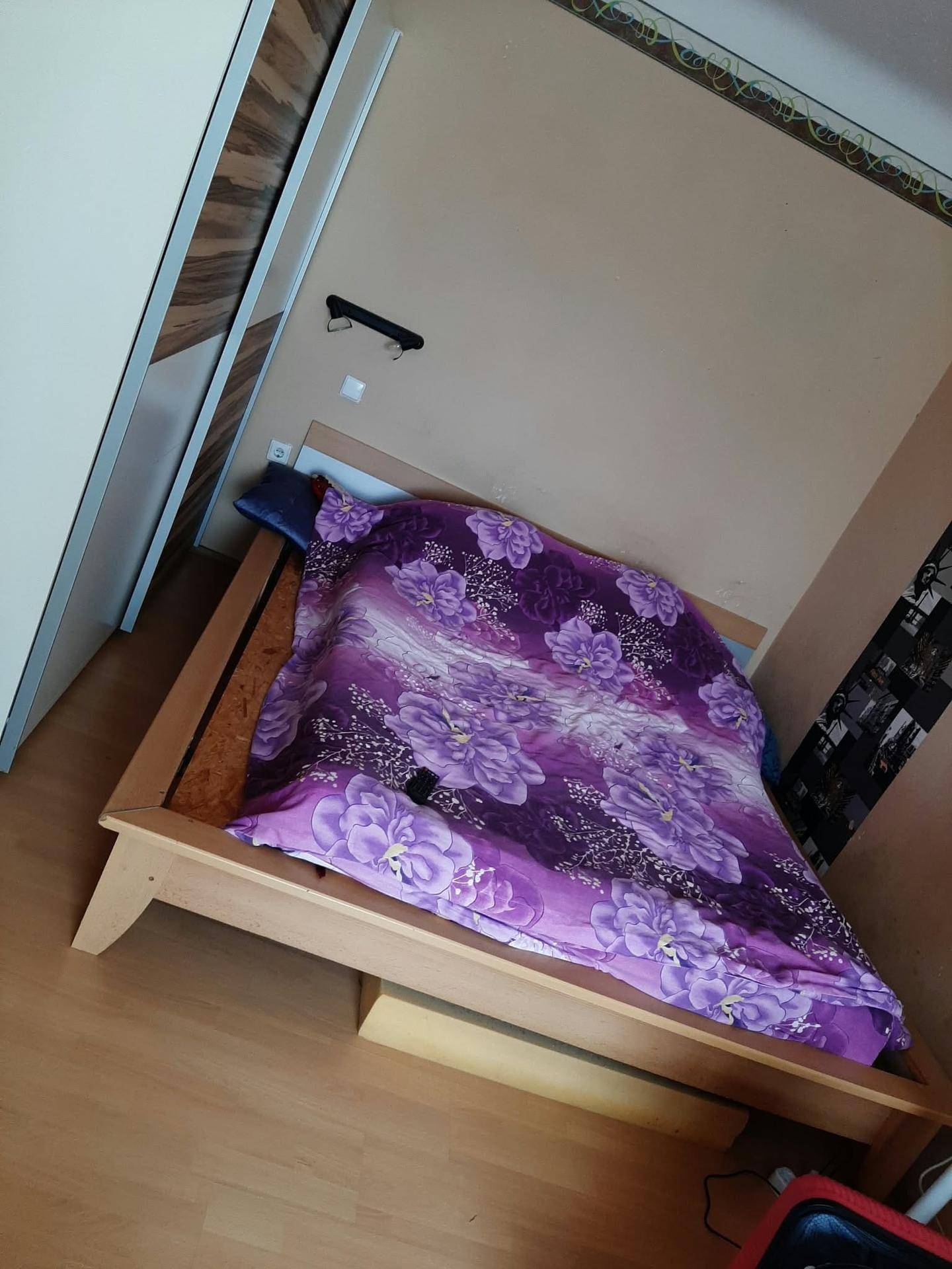 Schlafzimmer mit einfachem Holzbettgestell, lila Bettwäsche und hellem Laminatboden.