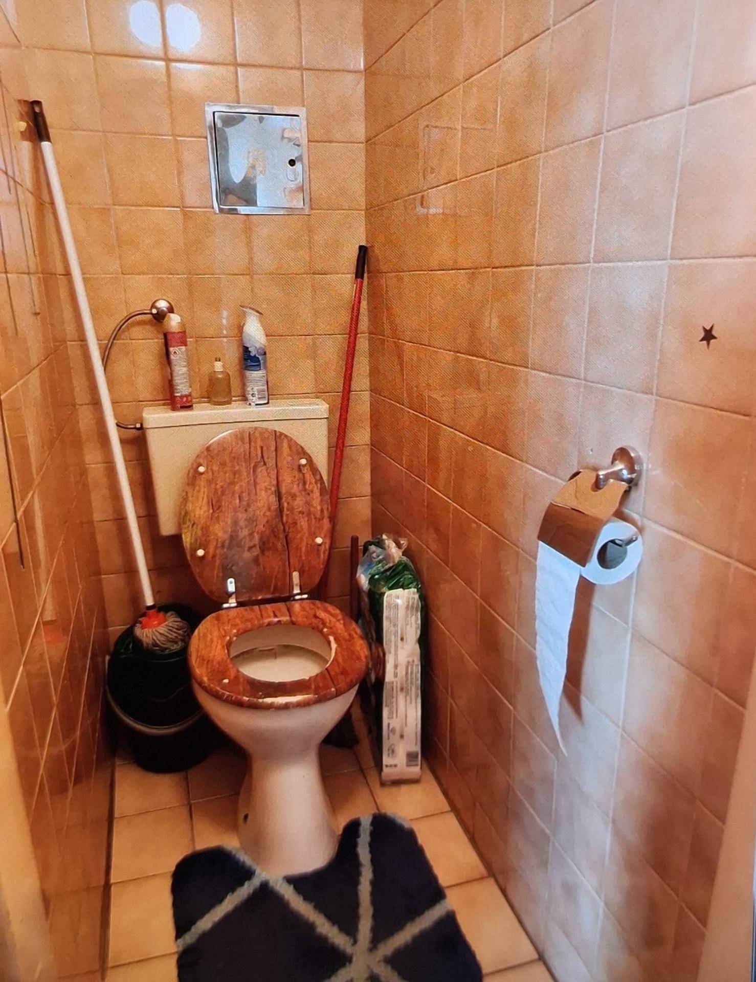 Separate Toilette mit braunen Fliesen und einem WC mit Holzdeckel im 80er-Jahre-Stil.