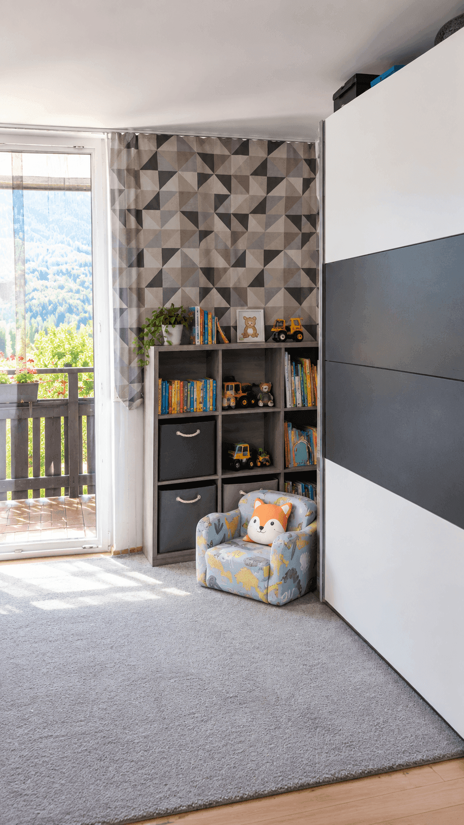 Helles Kinderzimmer mit Teppichboden, Bücherregal und Balkontür mit Bergblick.