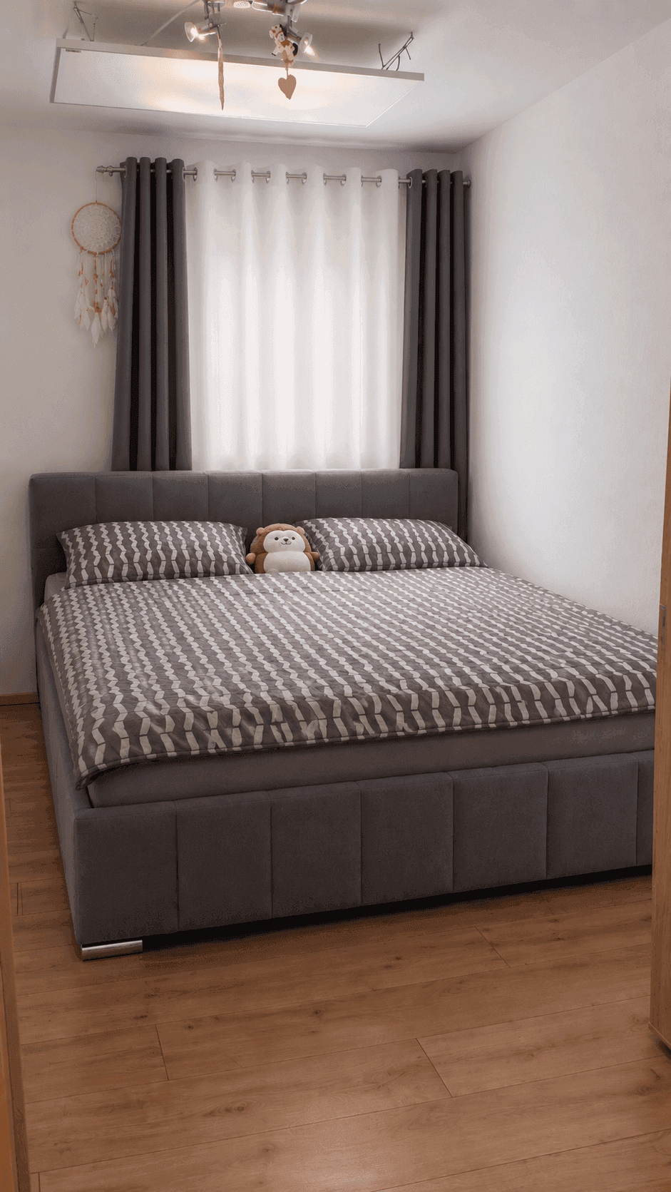 Schlafzimmer mit grauem Polsterbett, Holzboden und Vorhängen vor dem Fenster.