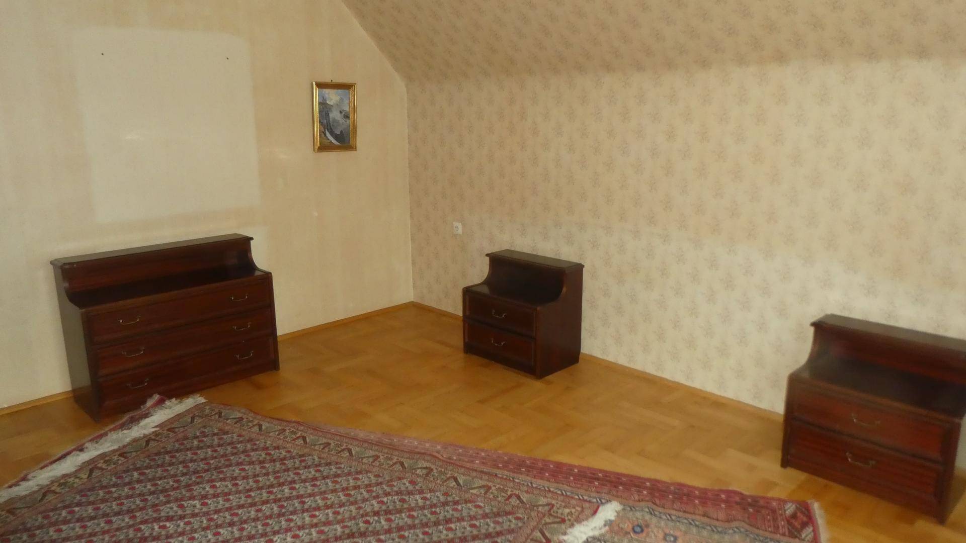 Schlafzimmer mit gemusterter Tapete, Holzmöbeln und einem Teppich auf dem Parkettboden.