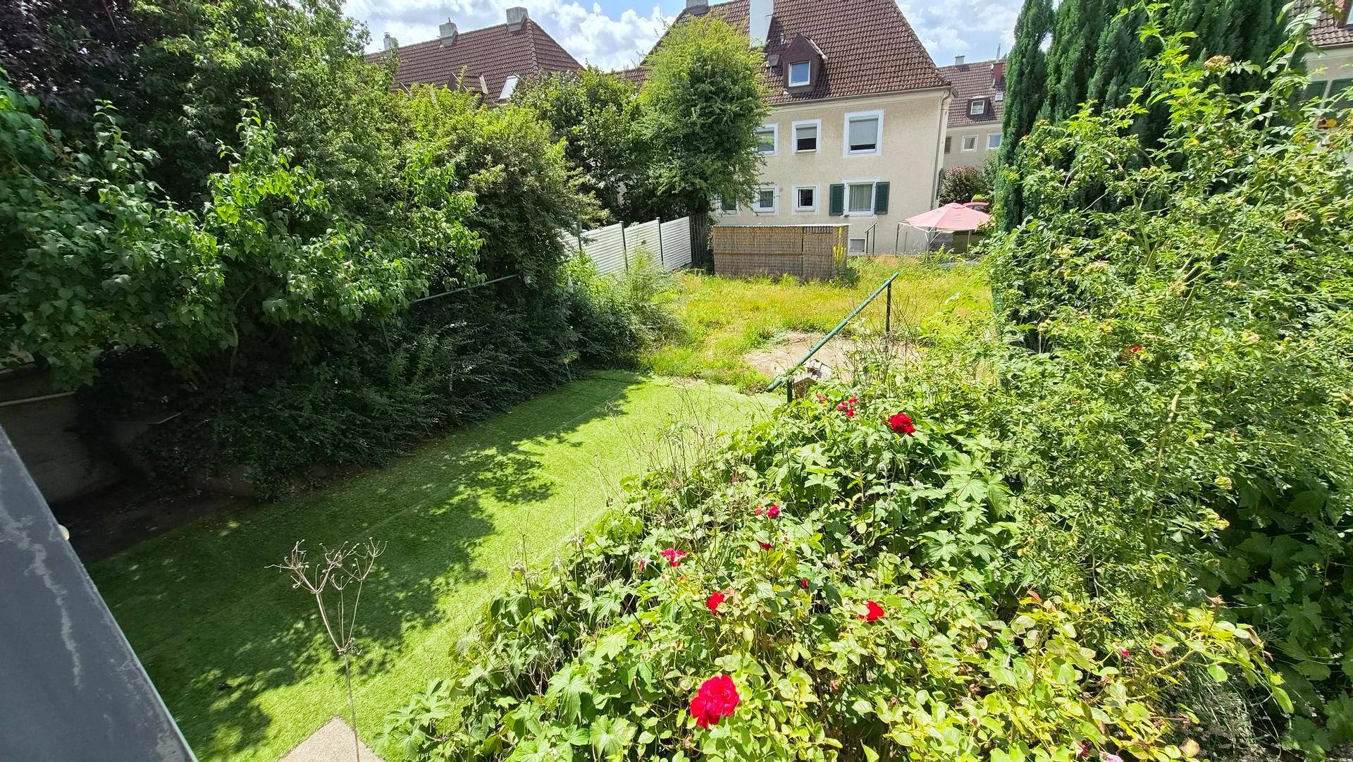 Blick auf den weitläufigen Gartenbereich mit viel Grün und angrenzenden Nachbargebäuden unter blauem Himmel.