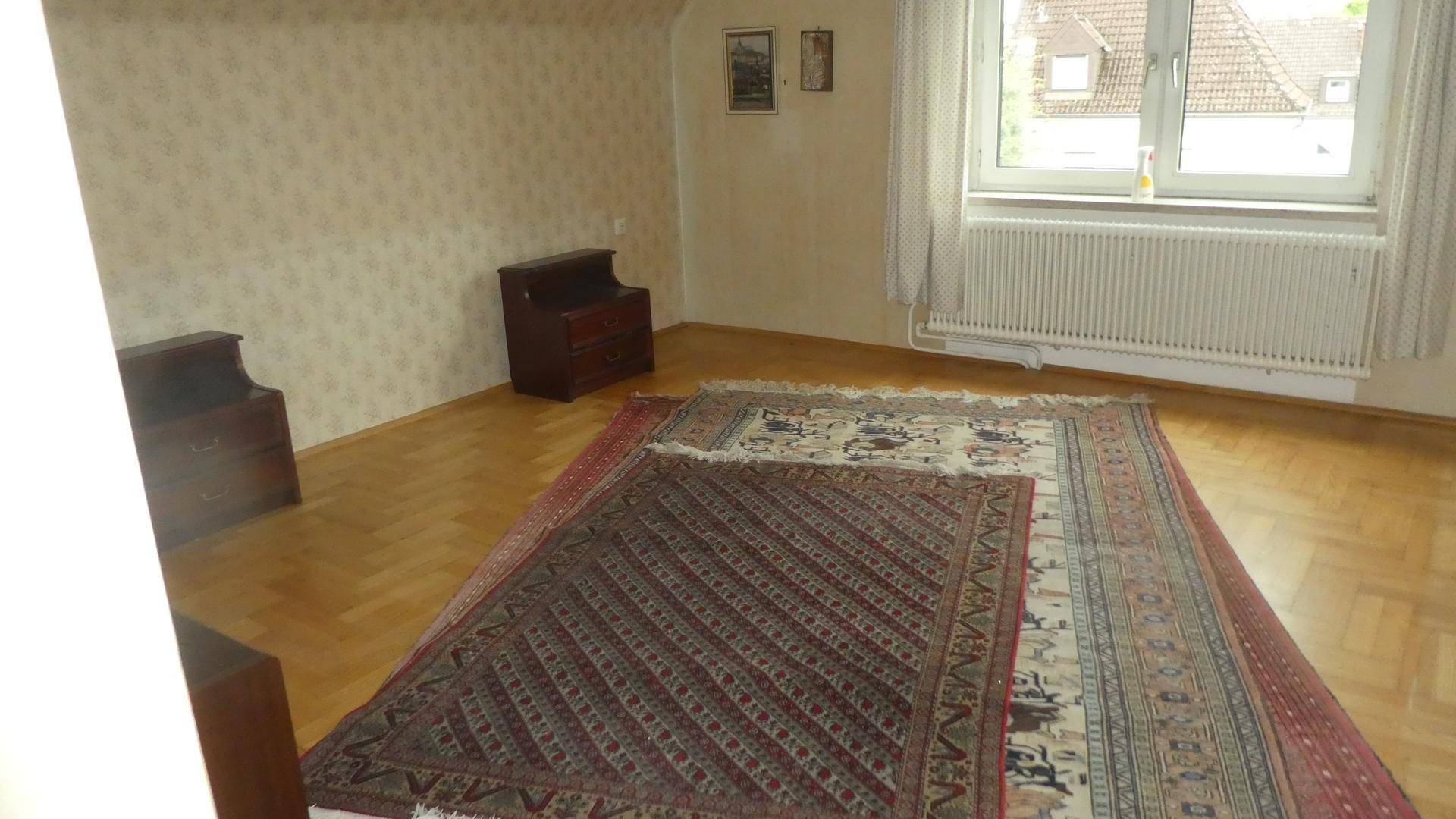 Helles Schlafzimmer mit Parkettboden, Teppich, Holzmöbeln und Fenster mit Blick ins Grüne.