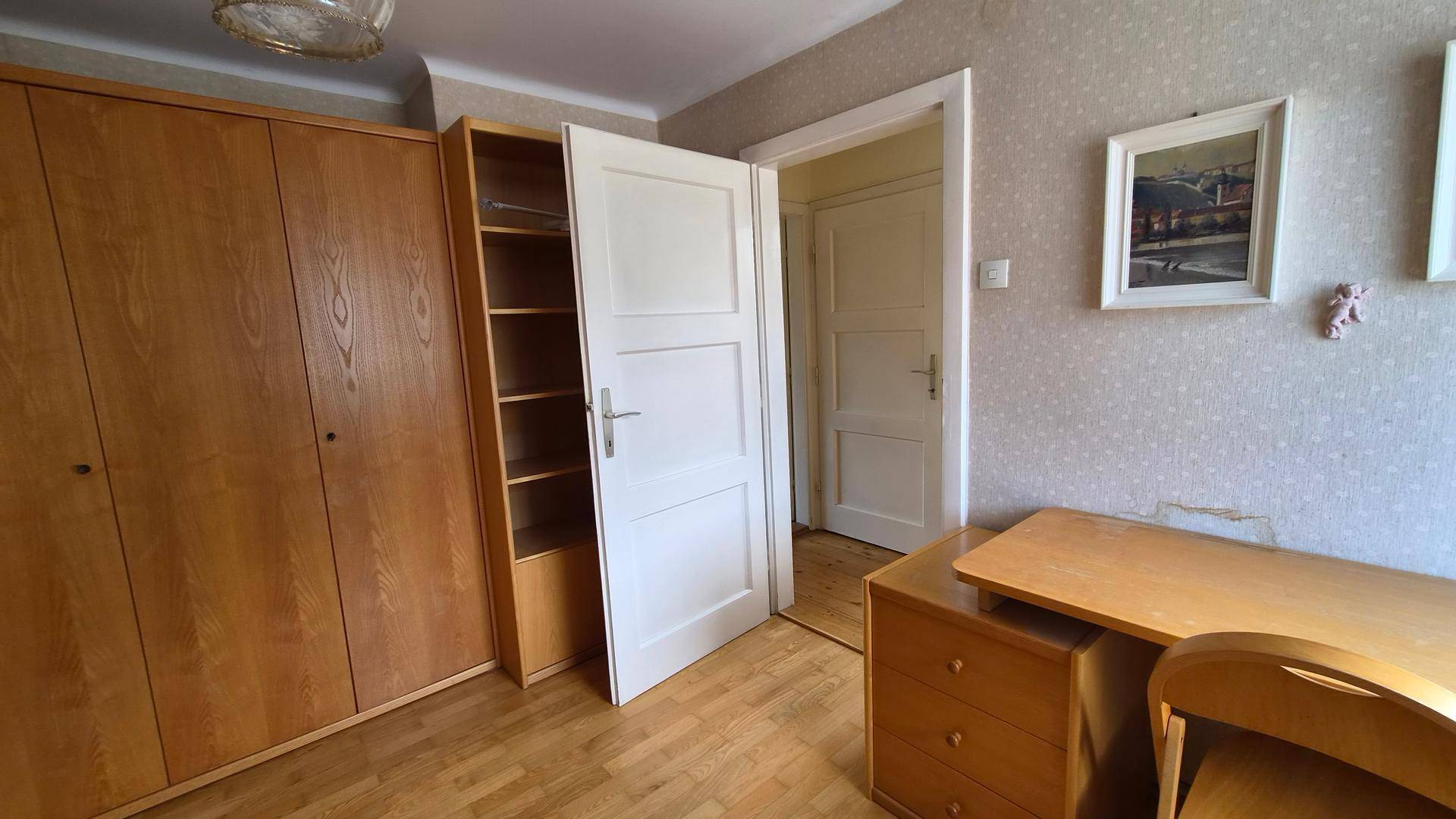 Schlafzimmer mit Holzschrank, Schreibtisch und Zugang zu einem weiteren Raum.