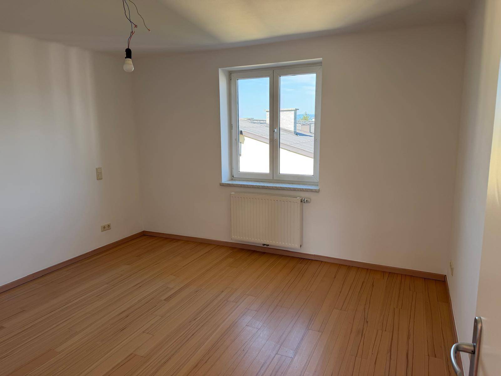 Lichtdurchflutetes, unmöbliertes Zimmer mit Holzboden und großem Fenster, bietet viel Tageslicht.