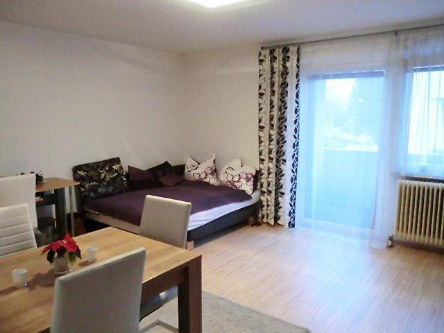 Heller Wohnbereich mit Esstisch, Schlafsofa und Vorhängen, die zum Balkon führen.
