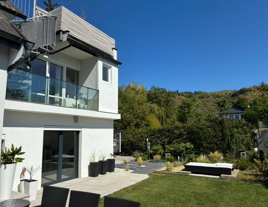 Ansicht des modernen Hauses vom Garten aus mit Balkon, Glasgeländer und üppiger grüner Umgebung.