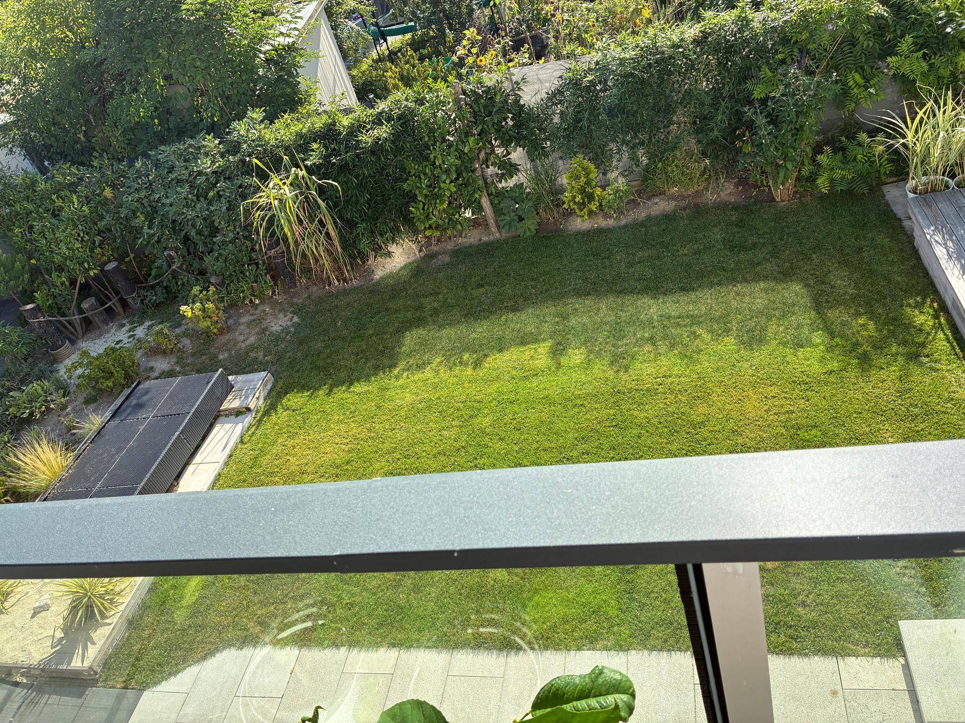 Blick von oben auf einen grünen Garten mit Rasenfläche, umgeben von Hecken und Bäumen, mit Solarpaneelen.