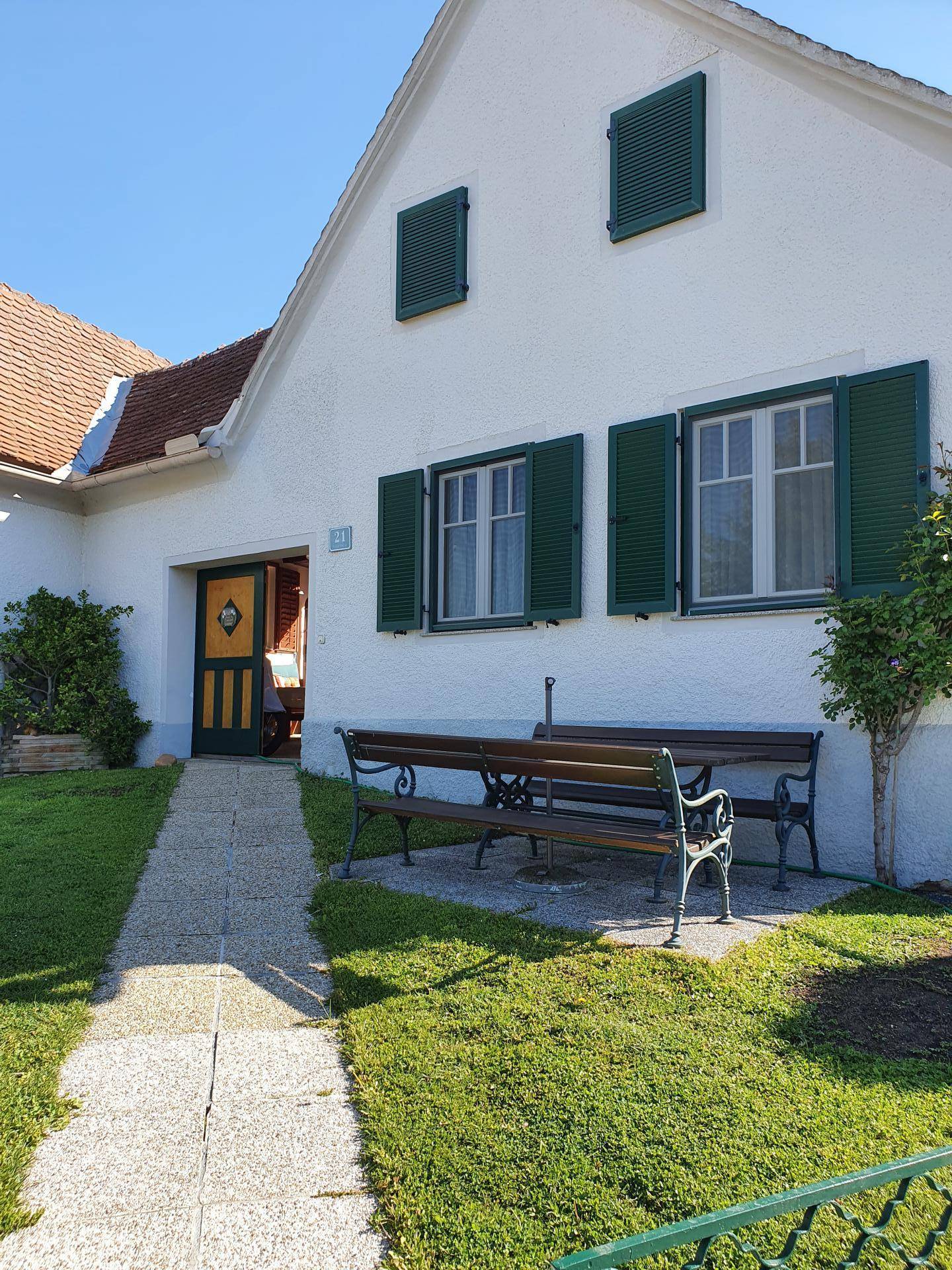 Weißes Einfamilienhaus mit grünen Fensterläden und einer Sitzbank im Vorgarten unter blauem Himmel.