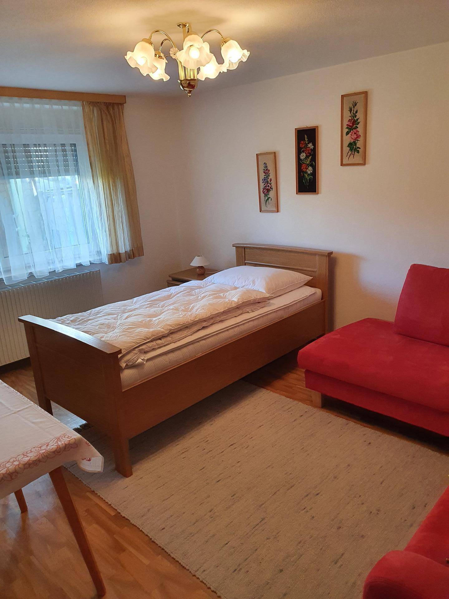 Helles Schlafzimmer mit Einzelbett, rotem Sofa und Holzboden, gut beleuchtet.
