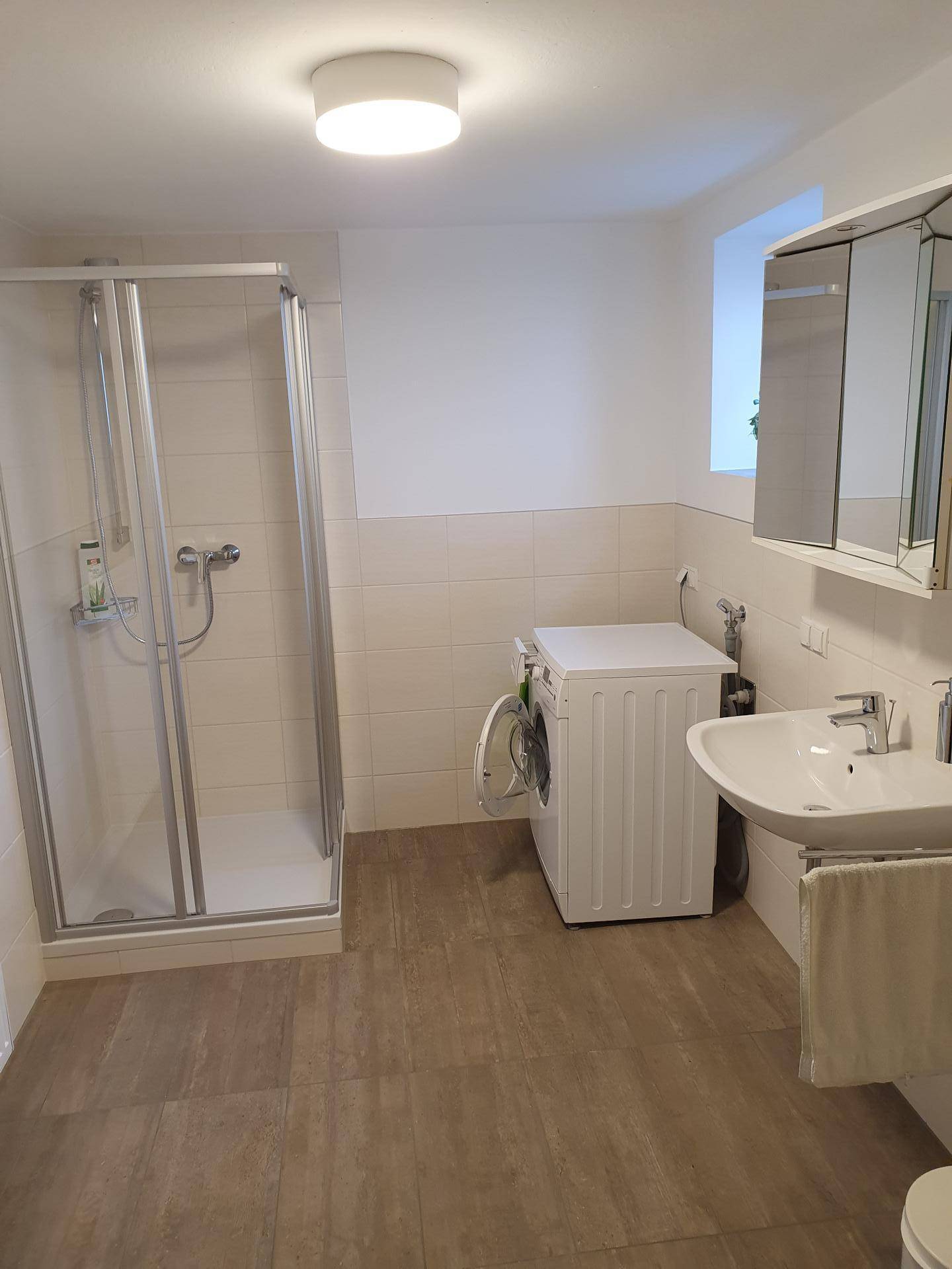 Modernes Badezimmer mit Duschkabine, Waschbecken und Waschmaschine, hell gefliest.