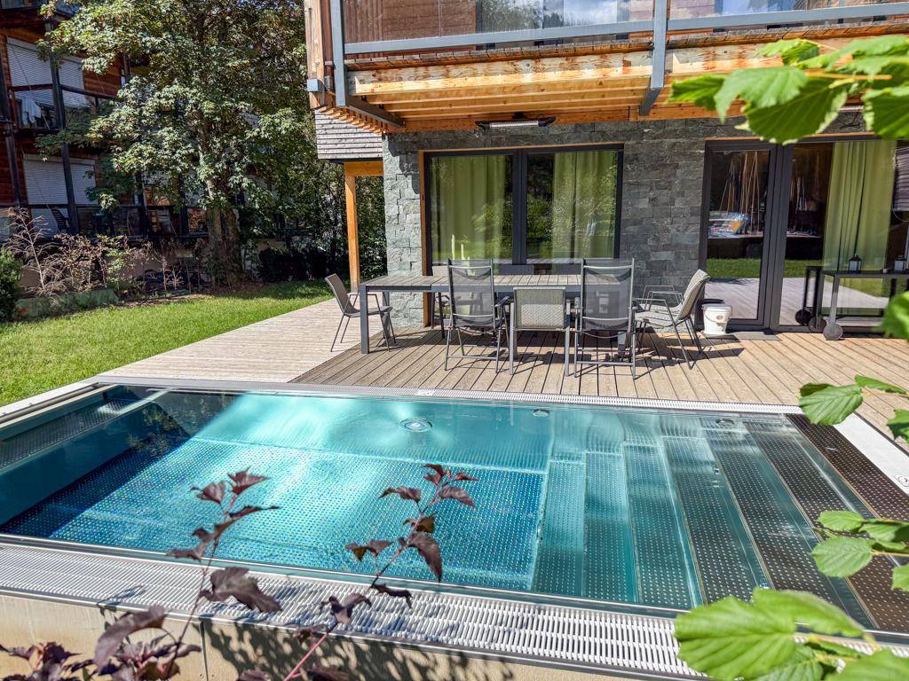 Sonnige Holzterrasse mit modernem Pool und Essbereich, umgeben von grüner Vegetation.