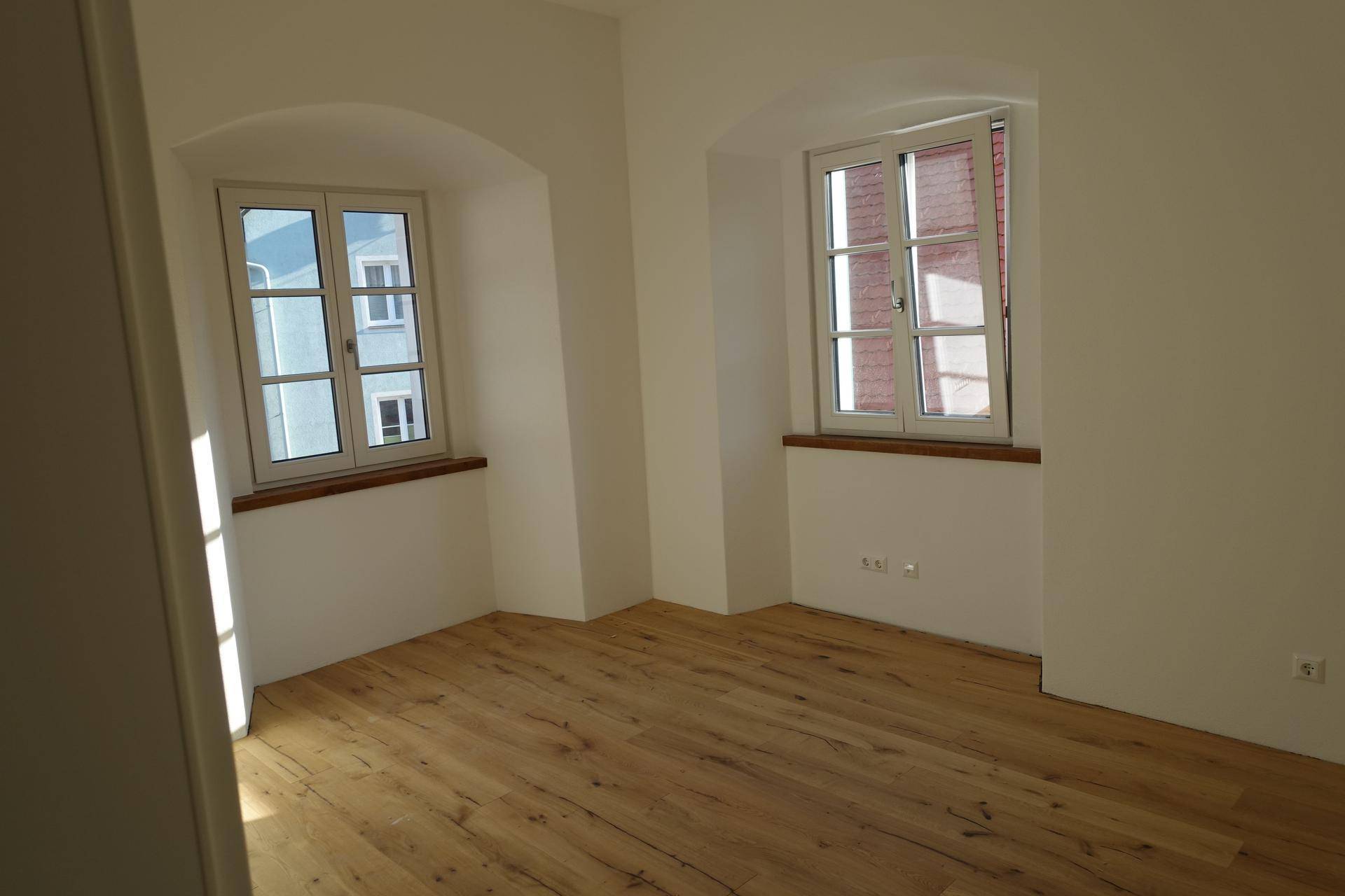 Heller, leerer Raum mit zwei Fenstern und warmem Holzfußboden.