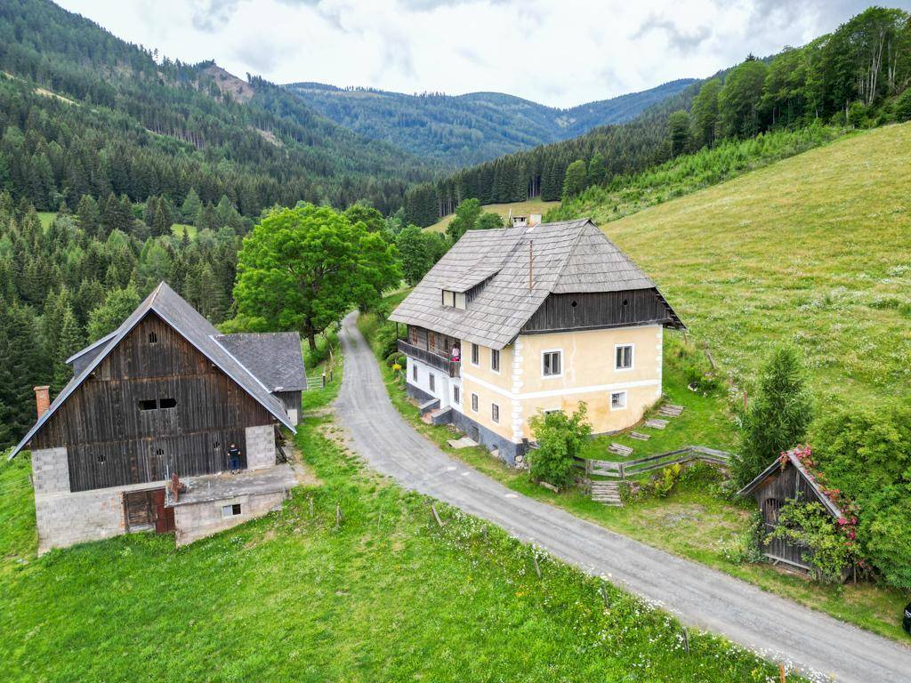 Luftaufnahme eines traditionellen Bauernhauses mit Nebengebäude, eingebettet in grüne Hügel und Wälder.
