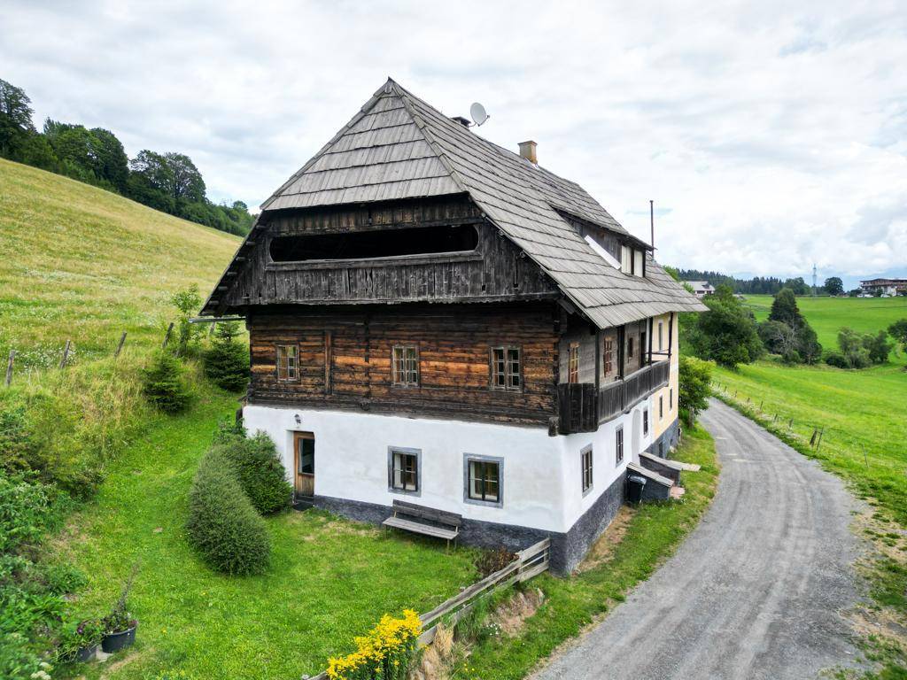 Historisches Bauernhaus mit Holzbalkon und Schindeldach, umgeben von grünen Hügeln und einem Feldweg.