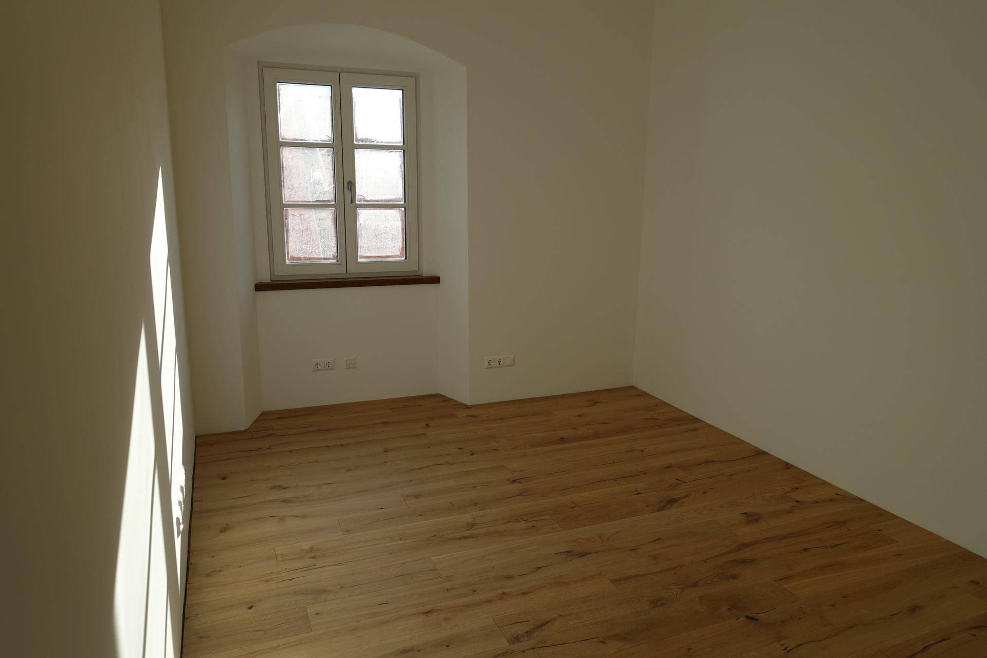 Sonniger, unmöblierter Raum mit einem Fenster und hellem Holzfußboden.