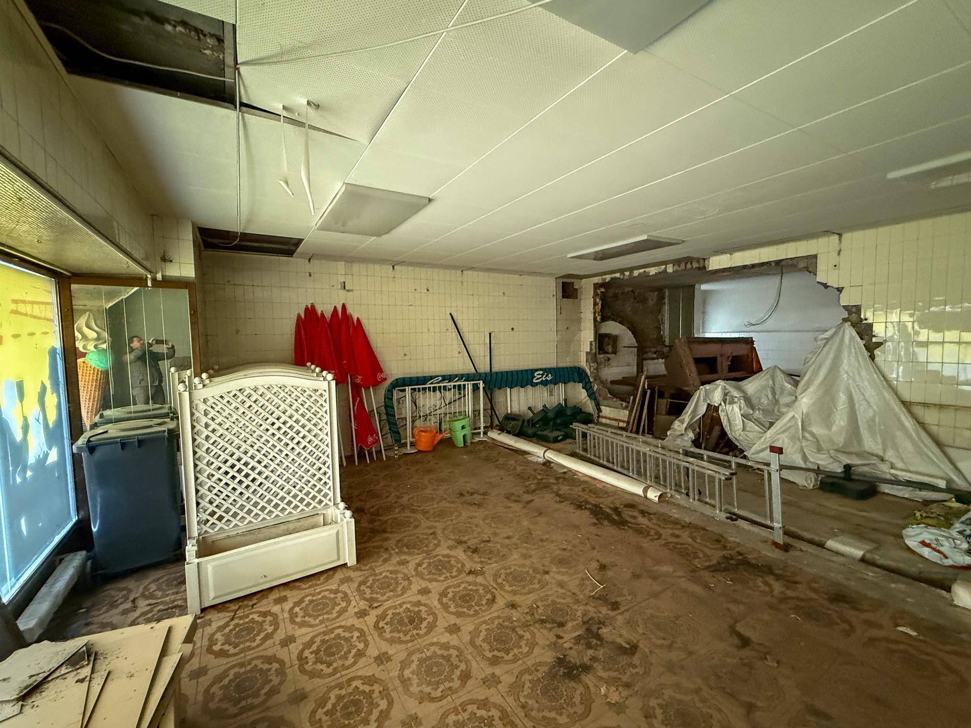 Weiterer Blick in den großen, leeren Raum mit Fliesenboden und sichtbaren Schäden an den Wänden.